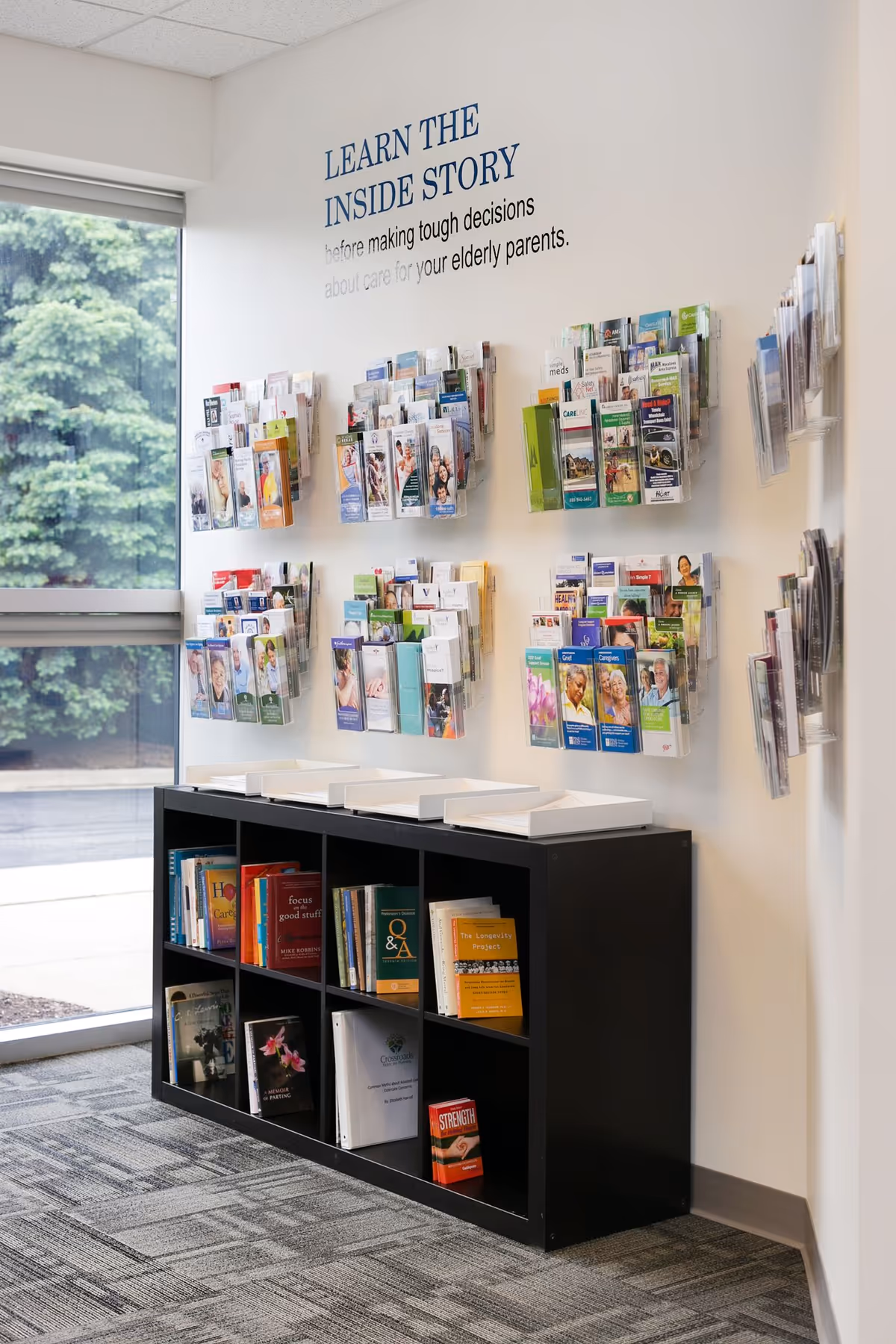 Interior brochure display with pamphlets on a wall near a window and a black bookshelf.