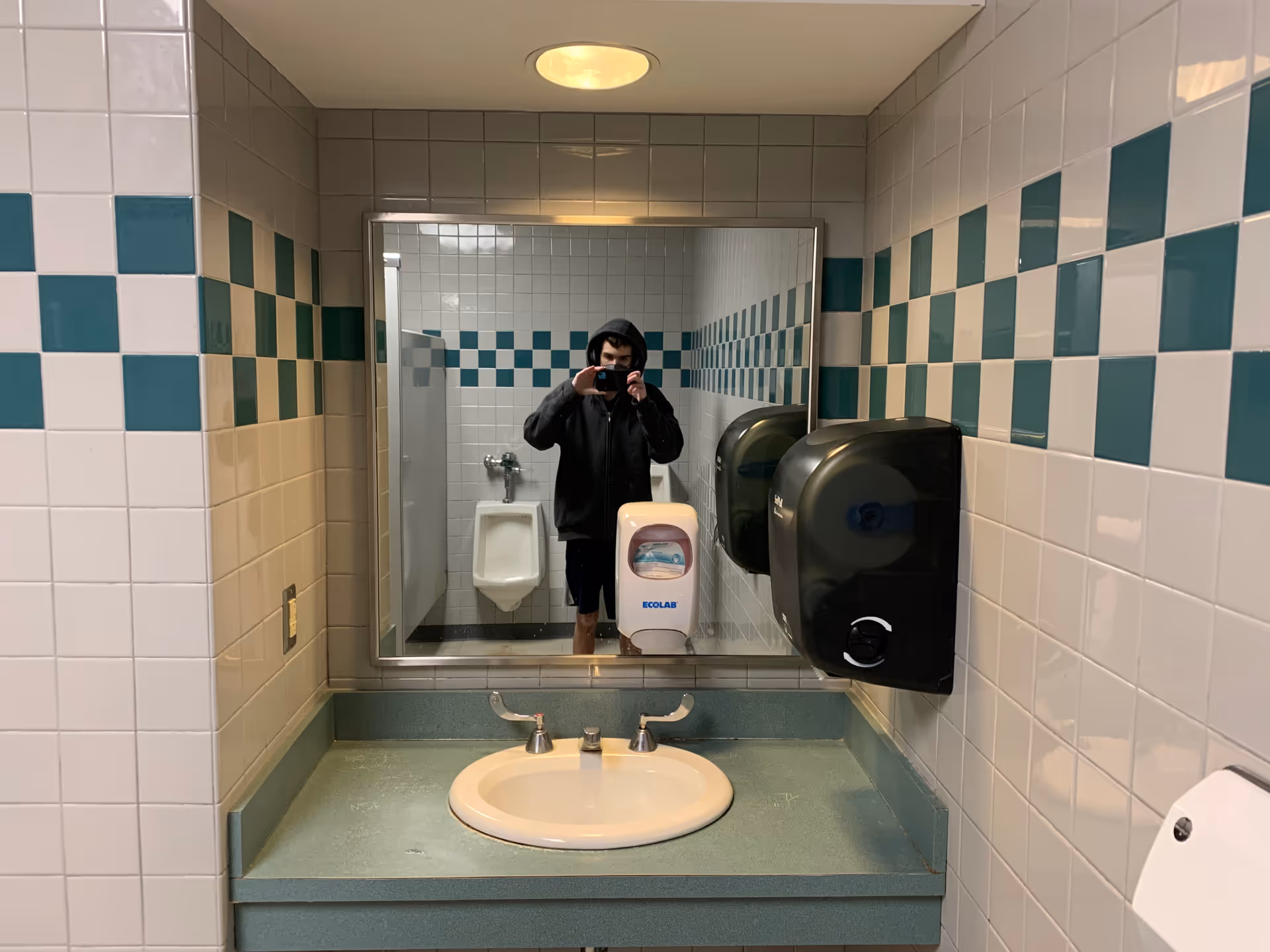 Person in hoodie takes a mirror selfie in a tiled bathroom with sink and dispenser.
