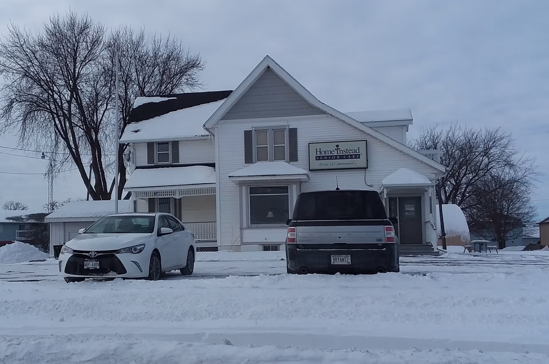 White two-story Home Instead Senior Care building with snow, parked cars in a winter lot.