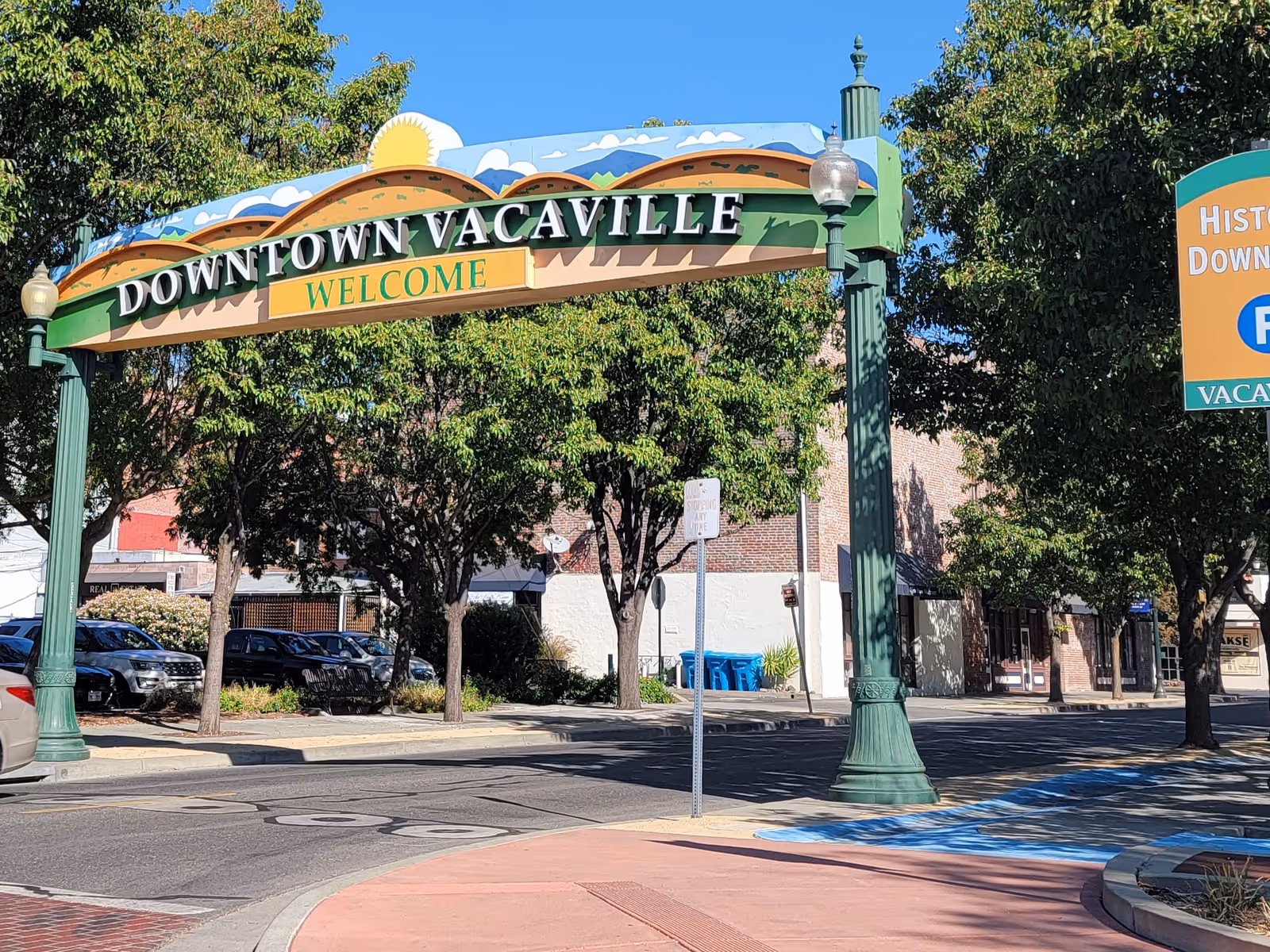 Colorful archway sign reading Downtown Vacaville Welcome spans a tree-lined street.
