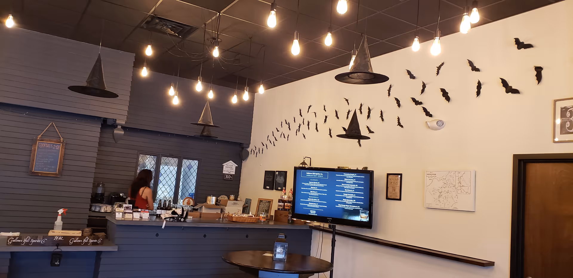 Cafe-style reception area with counter, hanging bulbs, TV, bat wall decor; a staff member behind counter.