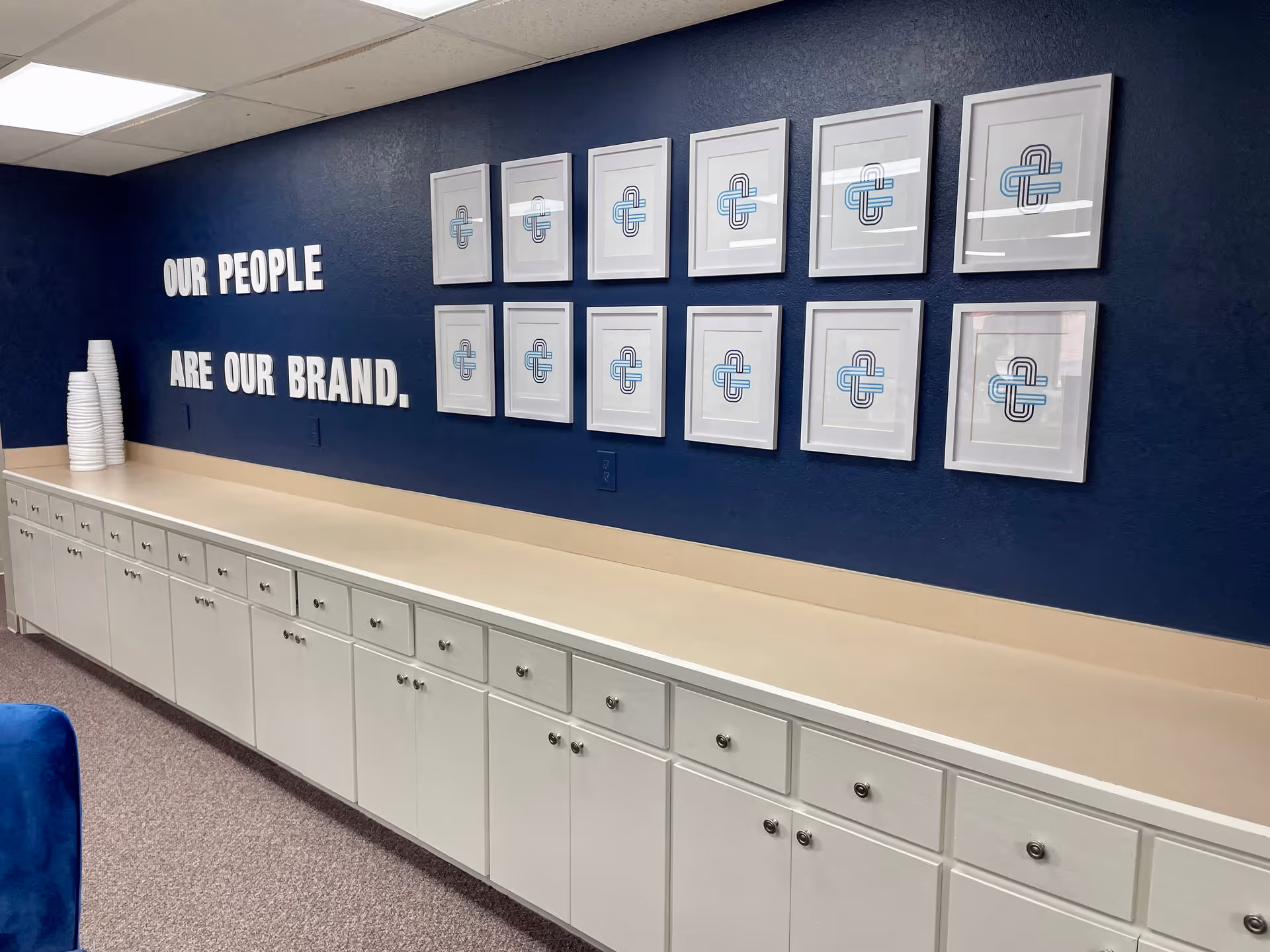 Blue wall featuring framed logos and the slogan Our People Are Our Brand above white cabinets.