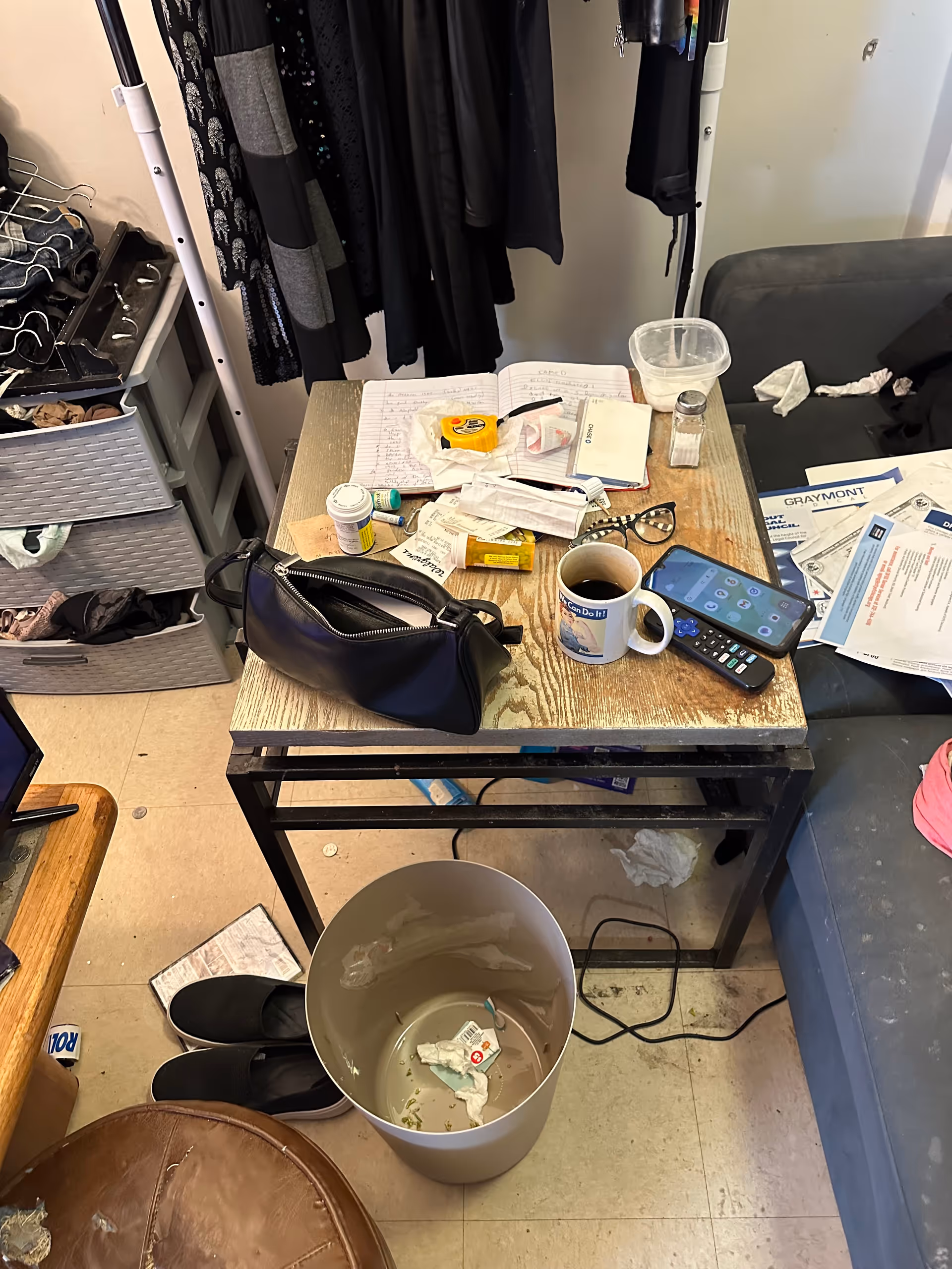Cluttered resident room desk with mug, phone, meds, papers, and clothing hanging nearby.