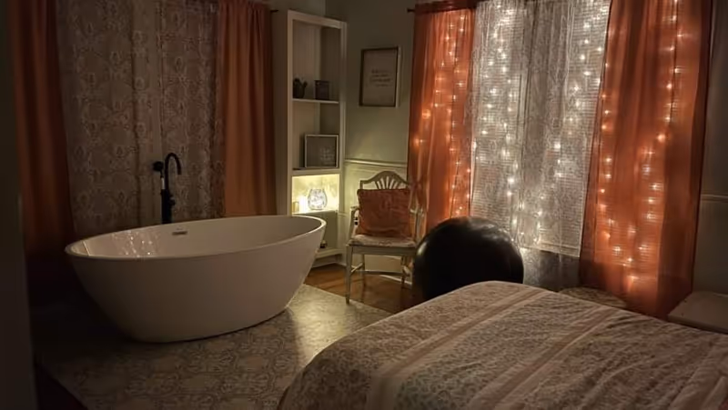 Dimly lit interior bedroom with a freestanding bathtub, bed, chair, and string lights by curtains.