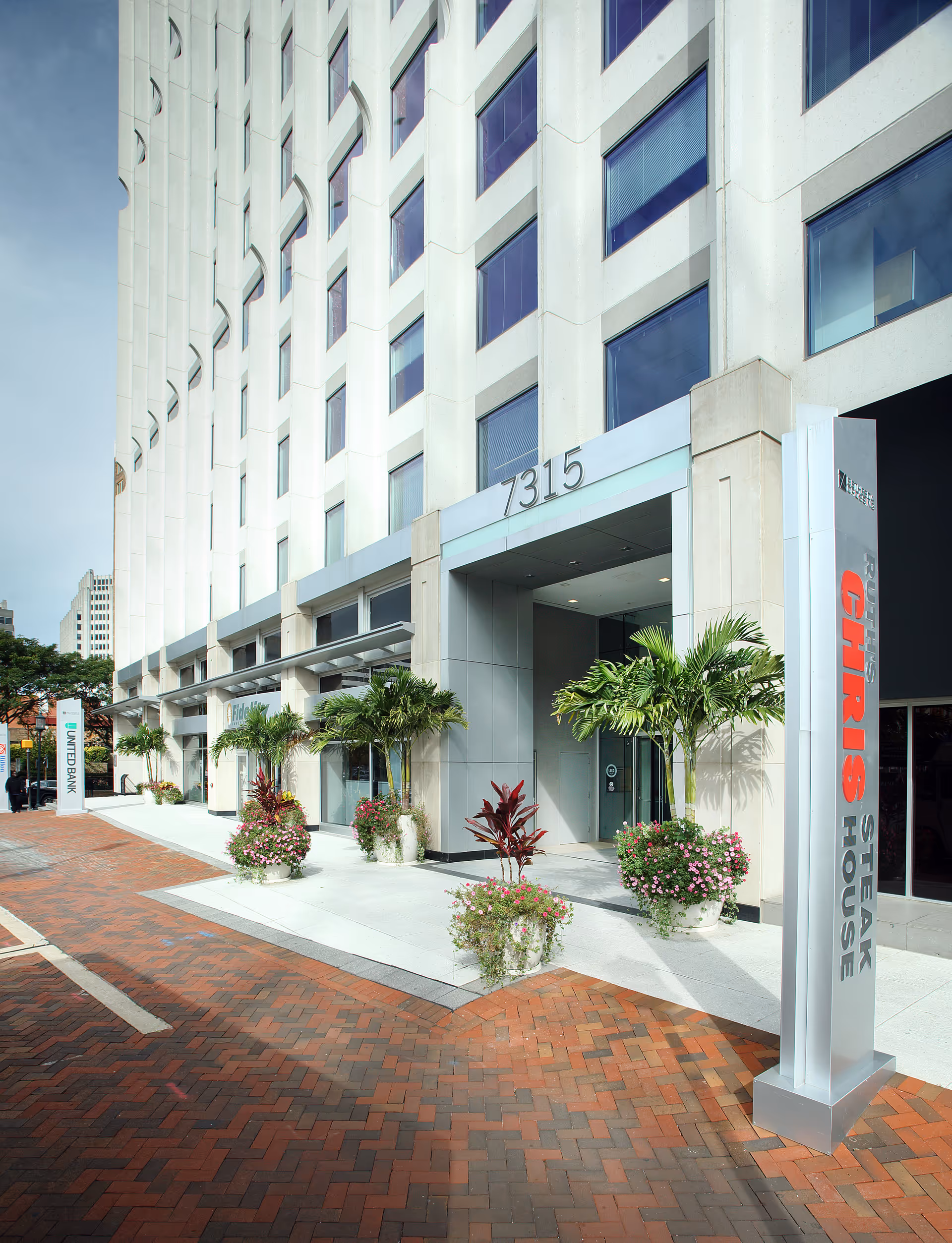 Exterior view of a modern multi-story building entrance with palm trees and planters.