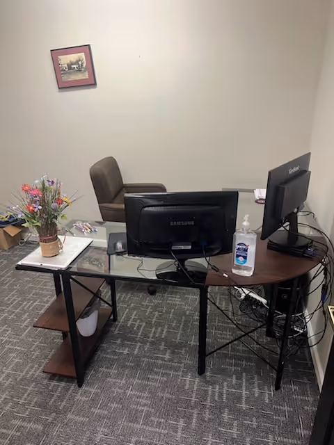 Reception-area office with dual computer monitors, sanitizer, and a flower arrangement on a glass desk.
