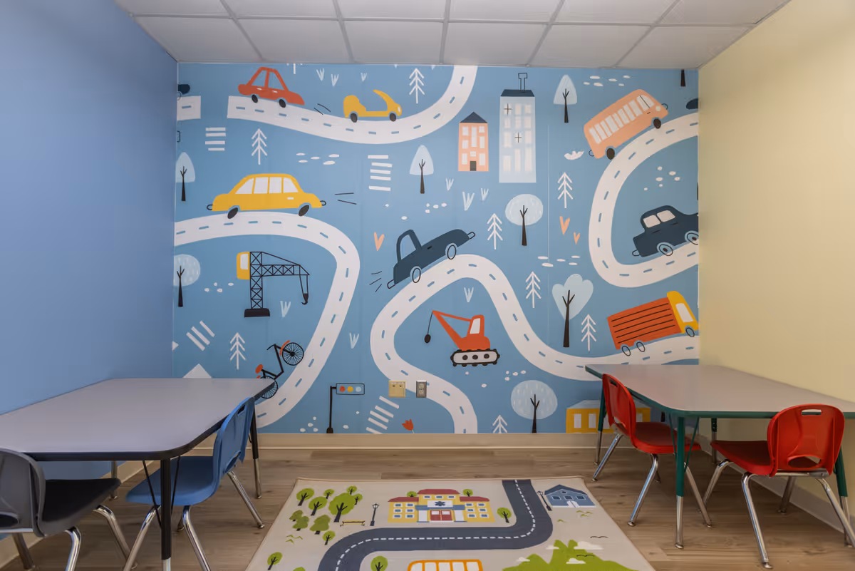 Bright children's activity room with a blue mural wall featuring roads and cars, tables and chairs.