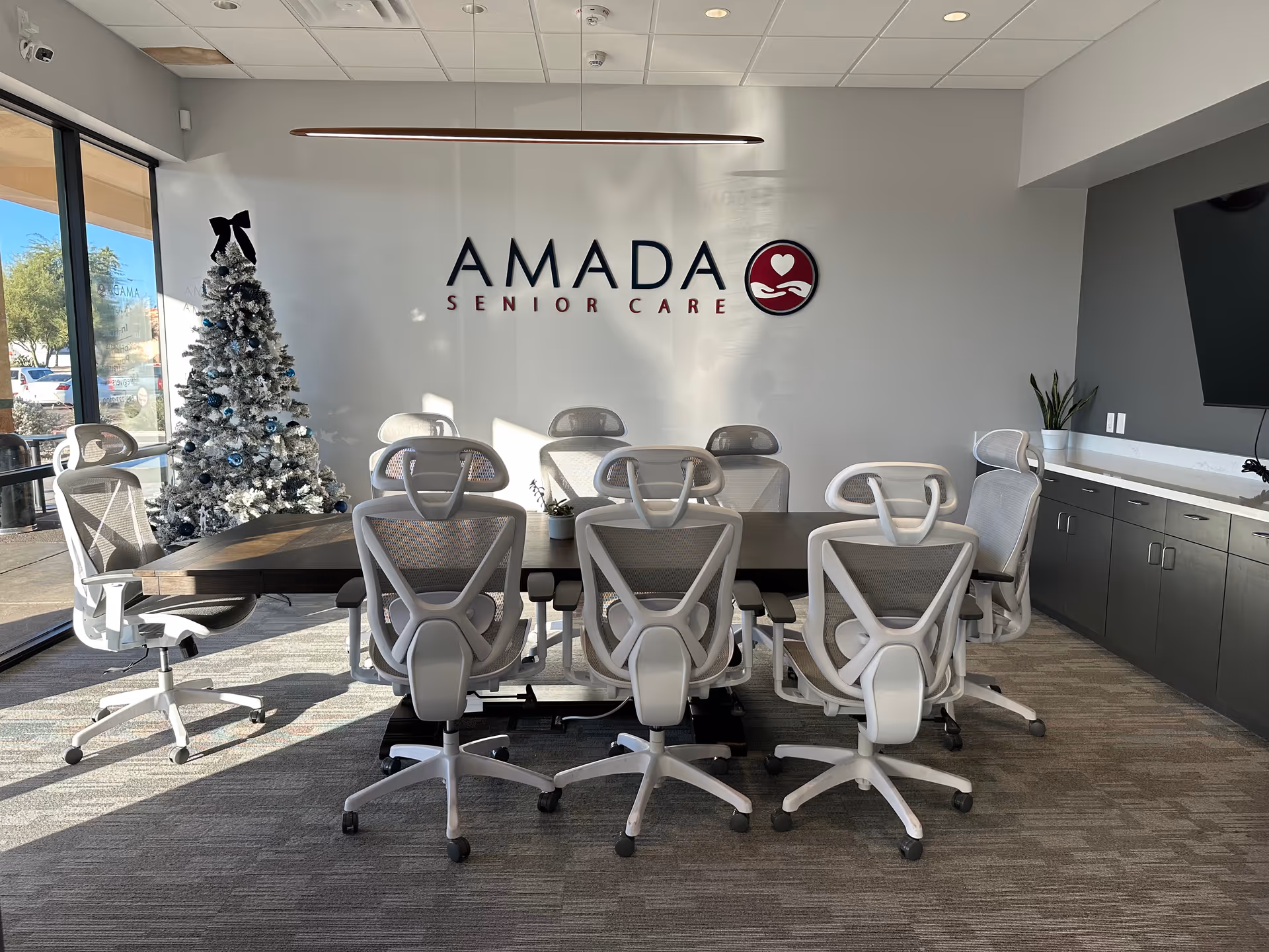 Modern Amada Senior Care conference room with a long table, white chairs, and a Christmas tree.