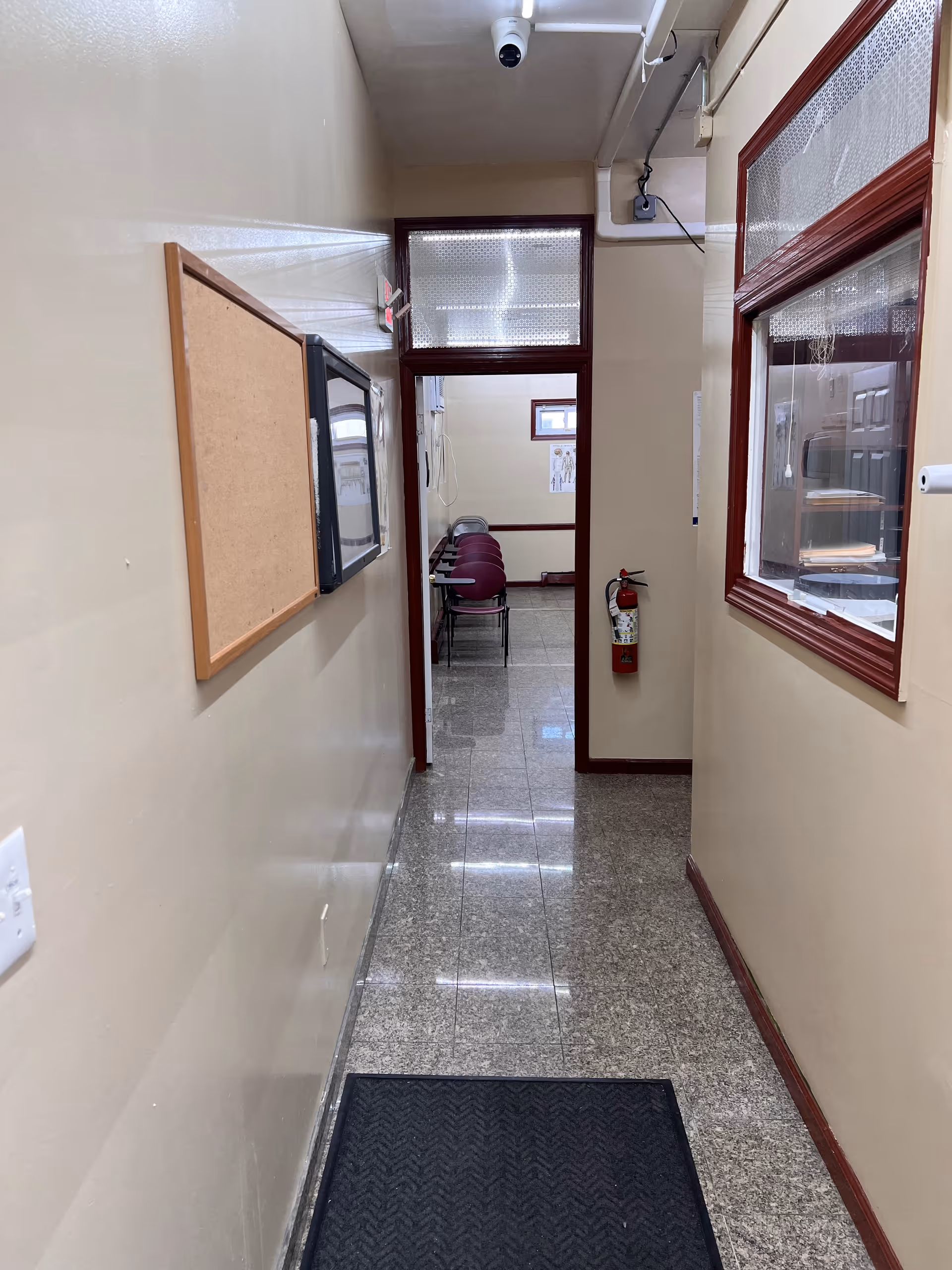 Narrow beige corridor leading to a small room with maroon chairs visible through a doorway.