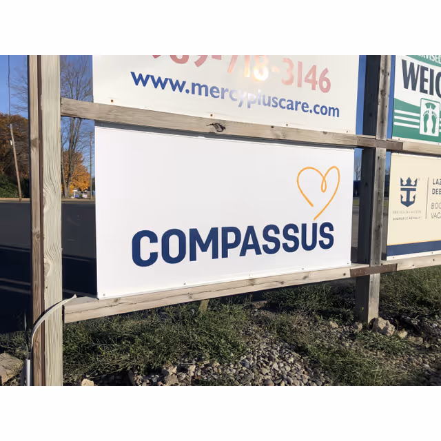 Exterior sign boards reading COMPASSUS mounted on a wooden frame outdoors.