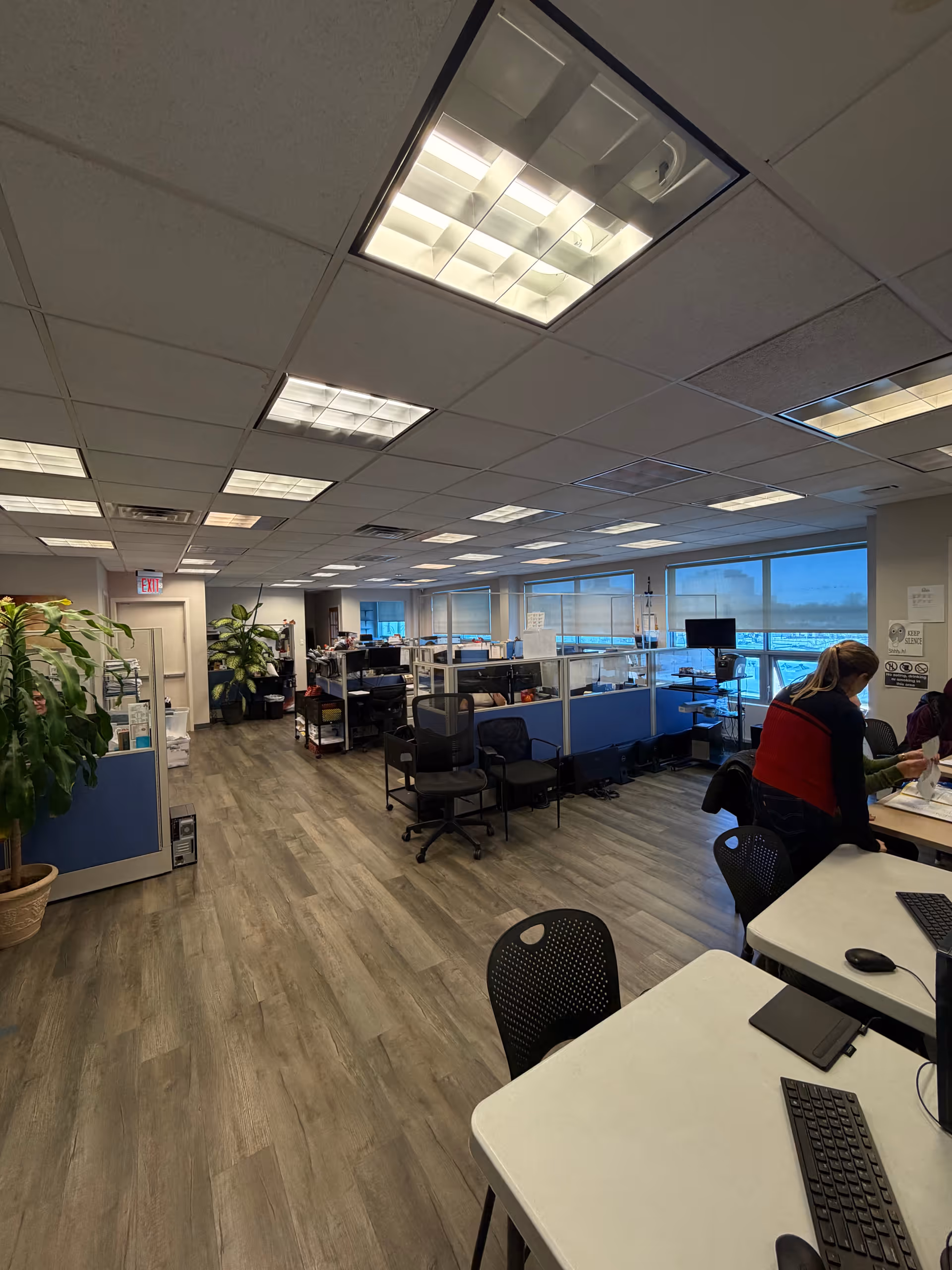 Open office area with cubicles, desks, computers, fluorescent lights, and a few staff members.