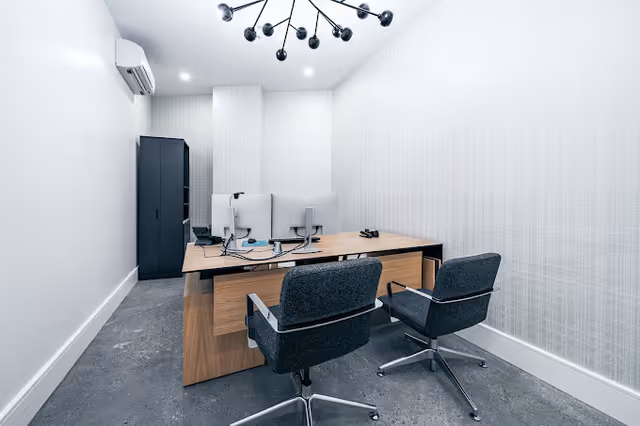 Minimalist office inside a white-walled room with a desk, chairs, and monitors.