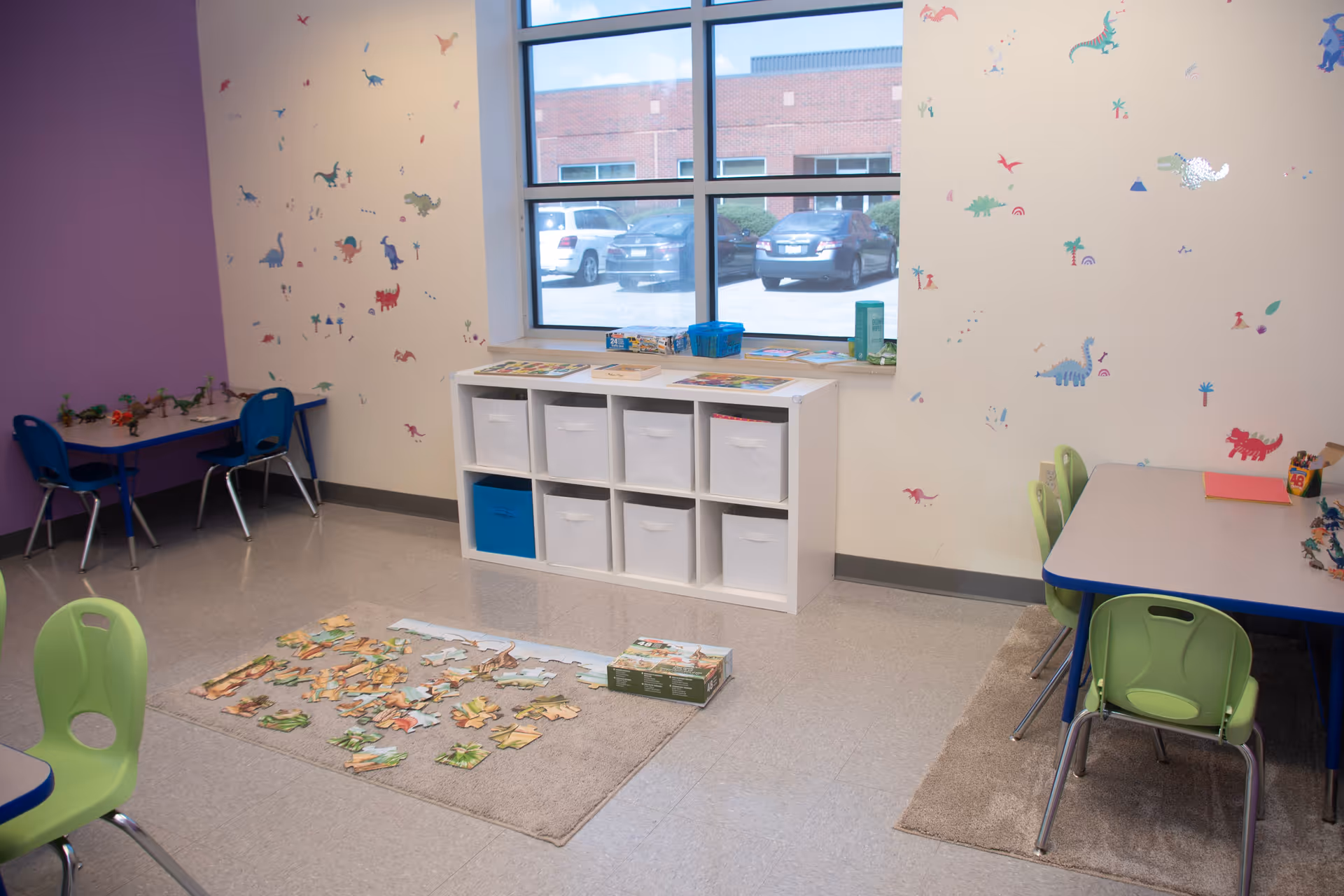Colorful interior playroom with tables, chairs, cubbies, and a puzzle rug by the window.