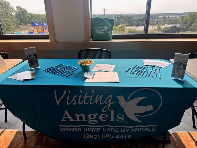 Indoor display table with Visiting Angels signage, brochures, and pens by a window.