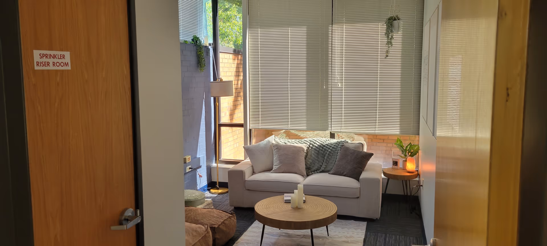 Cozy living room with beige sofa, round coffee table, cushions, plants, and blinds.