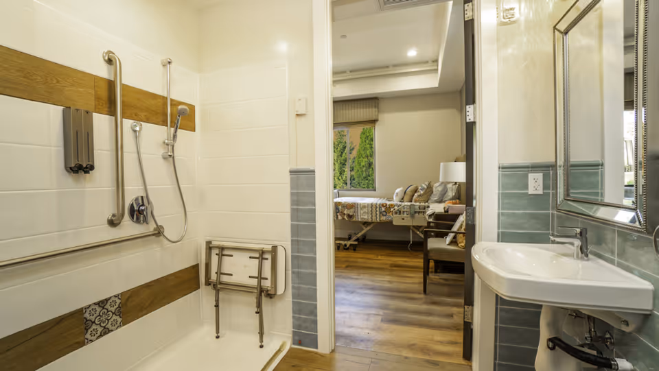 Accessible bathroom with grab bars, a shower chair, sink, and view into a resident room.