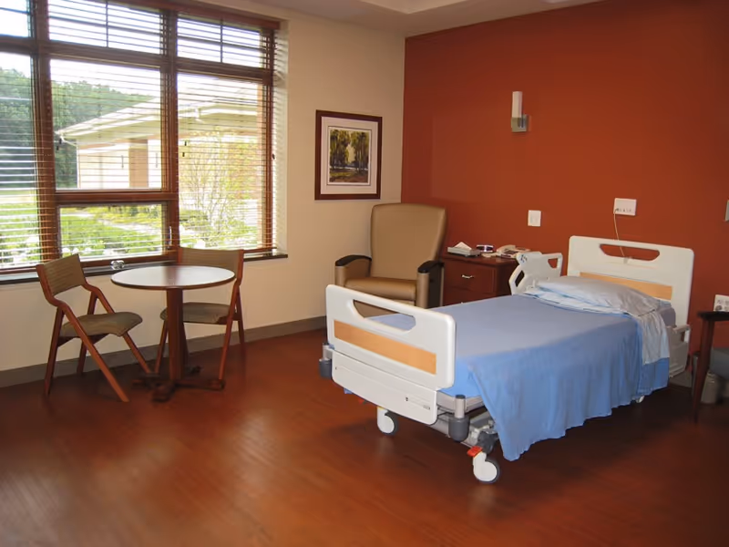 A patient bedroom in a senior living facility with a bed, chair, table, and window.