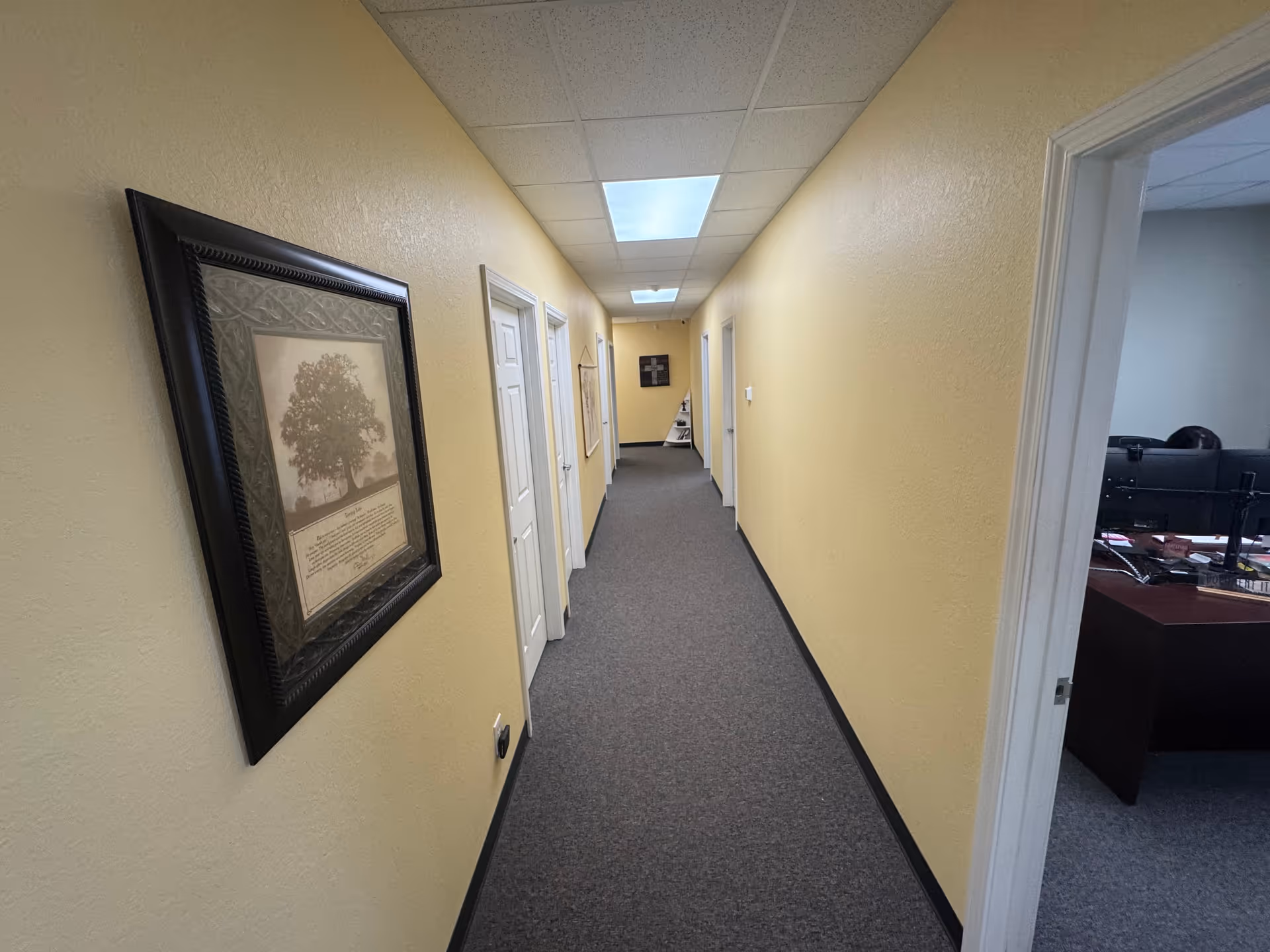 Narrow beige hallway in a senior living facility with doors, framed artwork, and an office.