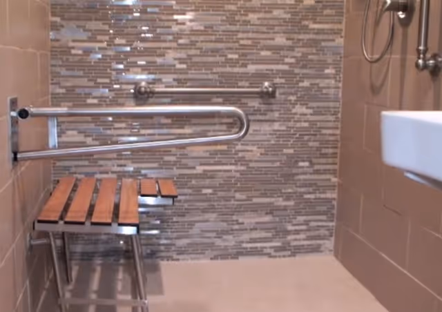 Accessible bathroom featuring grab bars, a wooden bench, and tiled wall.