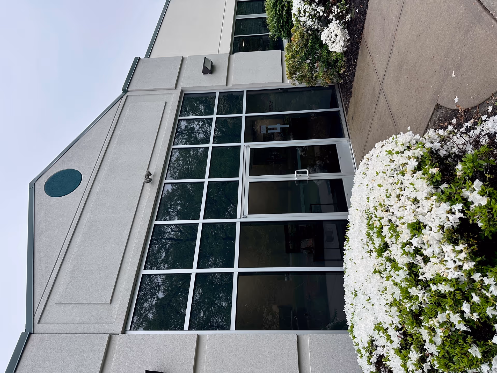 Exterior of a facility entrance with large glass doors and white flowering shrubs.