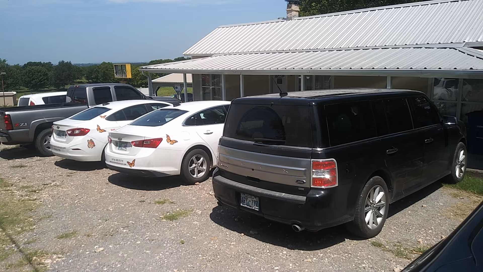 Gravel parking lot with several parked cars in front of a metal-roofed building.