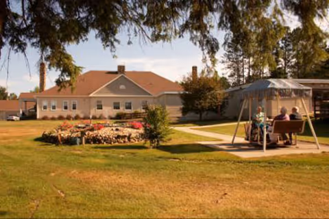 Senior living facility exterior with lawn, flower beds, and outdoor gazebo seating.