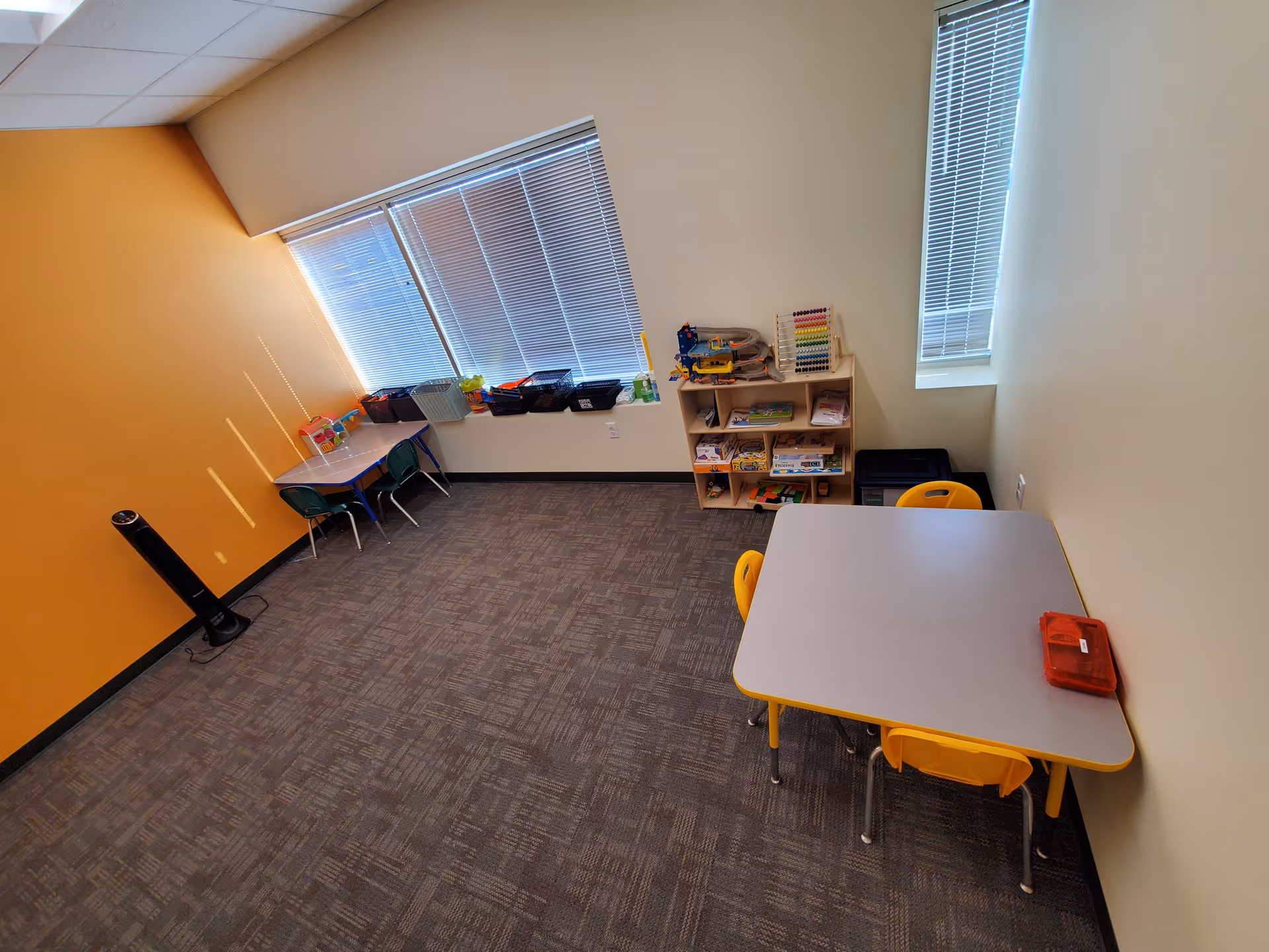 Small brightly colored classroom with tables, chairs, and shelves inside a facility.