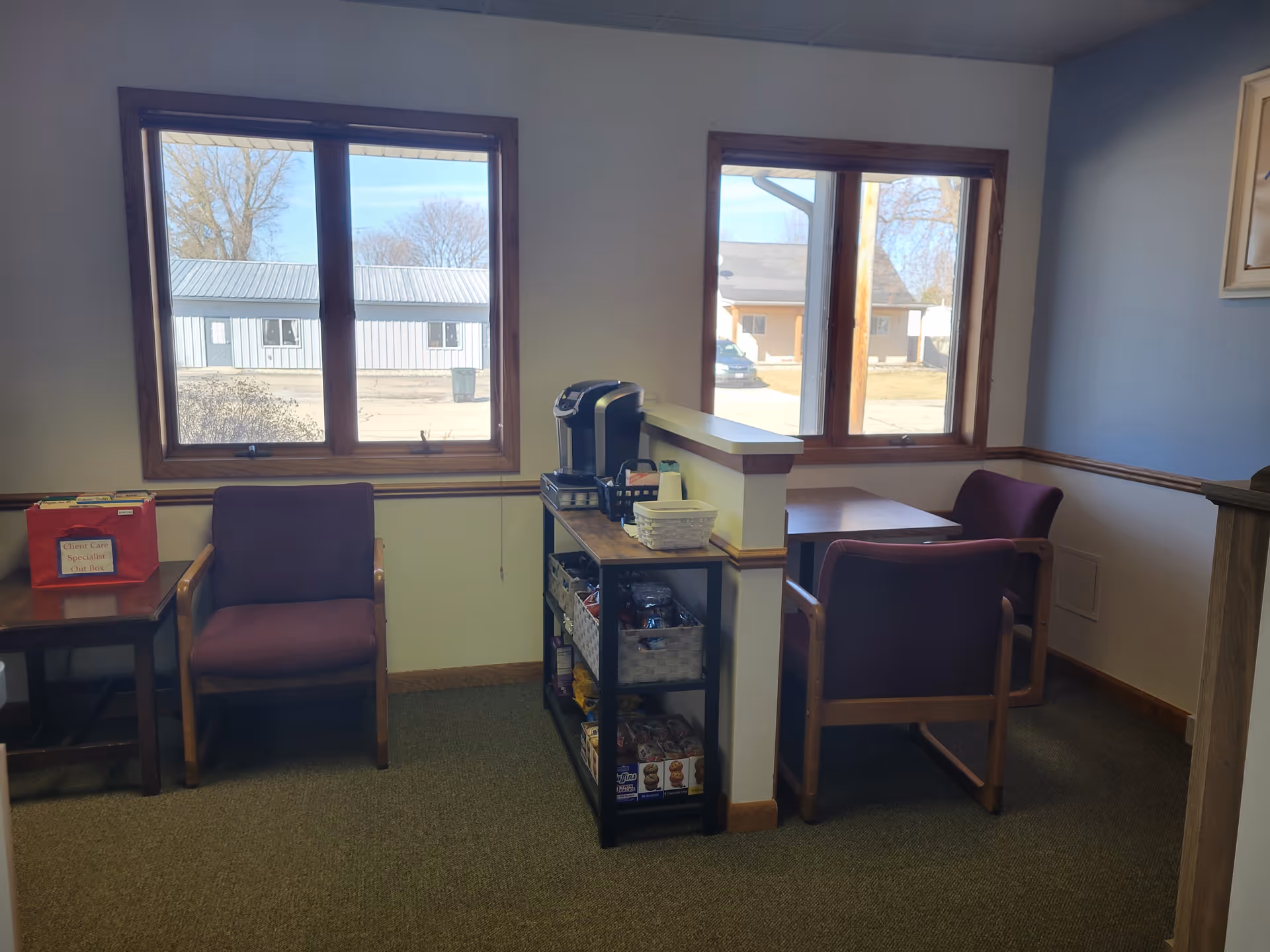 Small lounge area with burgundy chairs, a coffee station, and two windows.