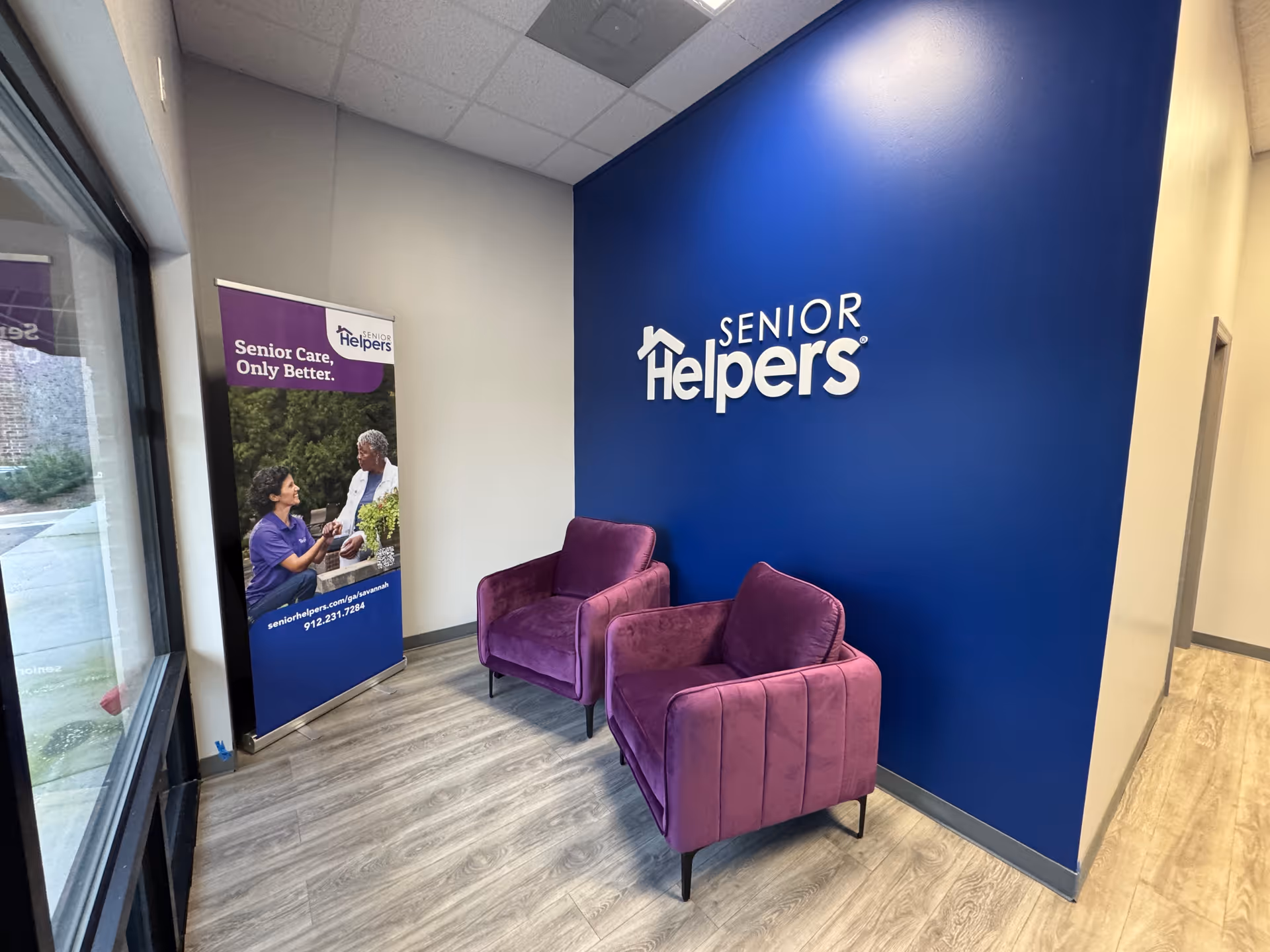 Blue wall with Senior Helpers logo, two purple armchairs, and a promotional banner.