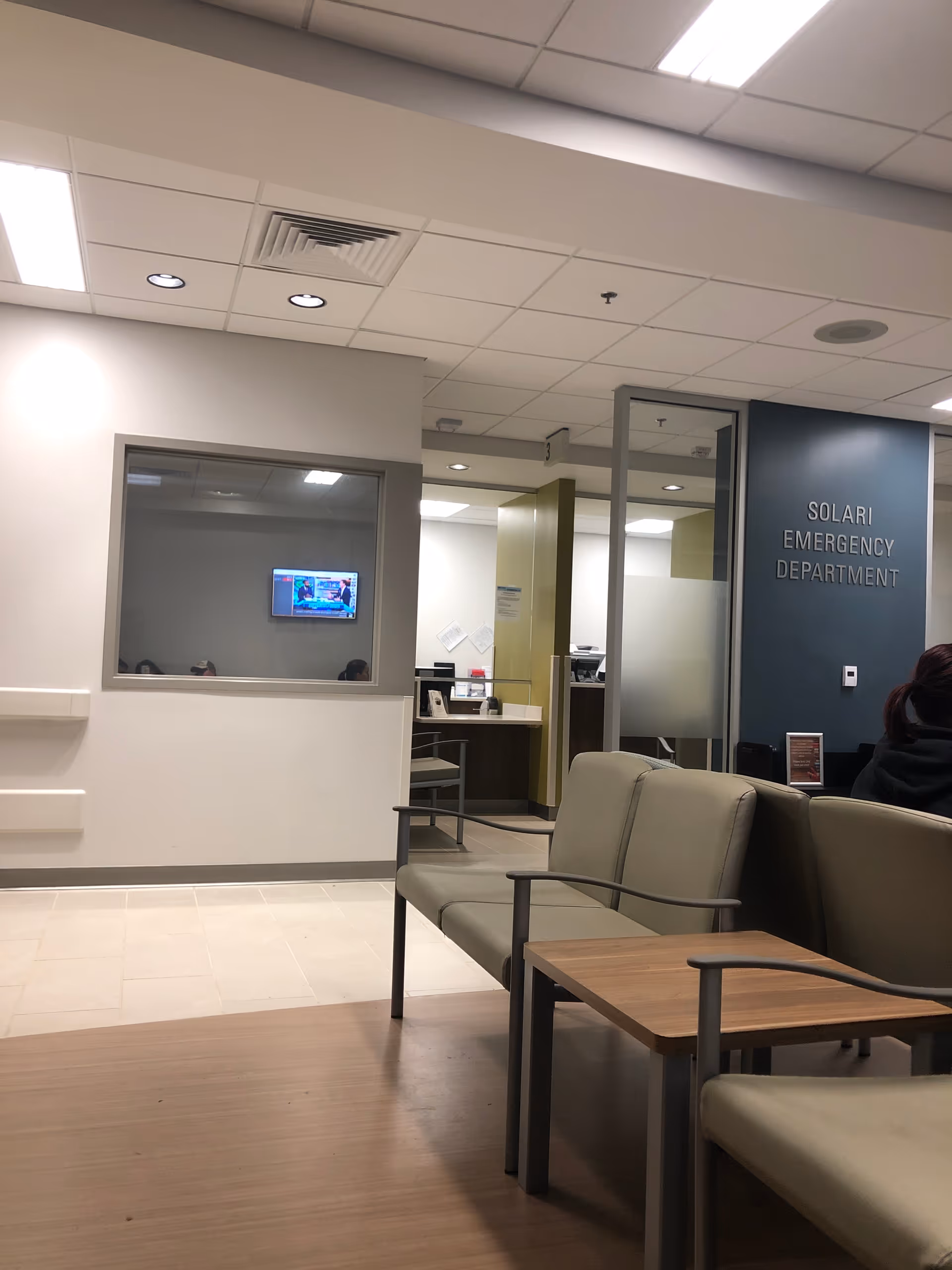 Hospital waiting area with beige chairs, a wooden table, and a view into a reception area.