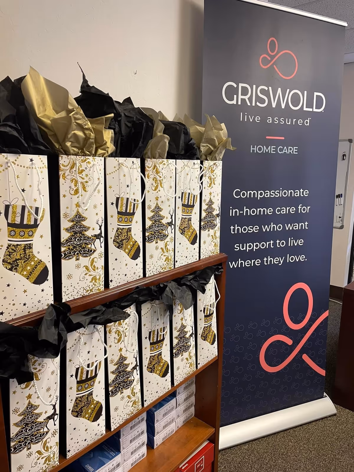 Decorated gift bags on shelves beside a Griswold Home Care banner in a facility interior.