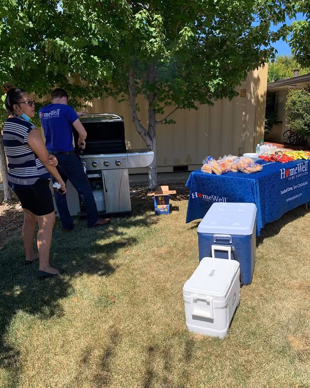 Outdoor gathering with a grill, a snack table with a HomeWell banner, and coolers.