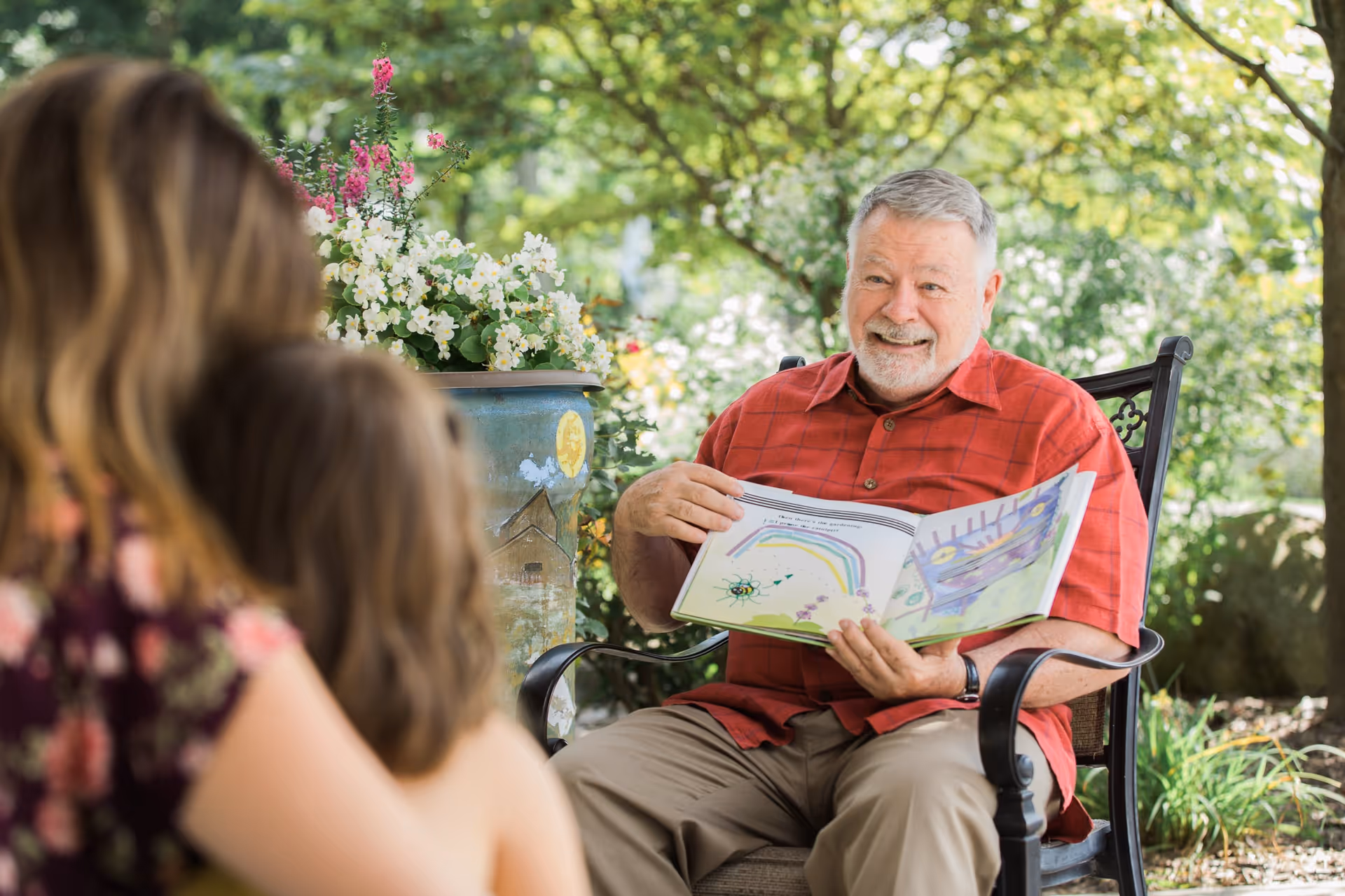 Senior man in a red shirt reads a children's book outdoors while seated in a chair.