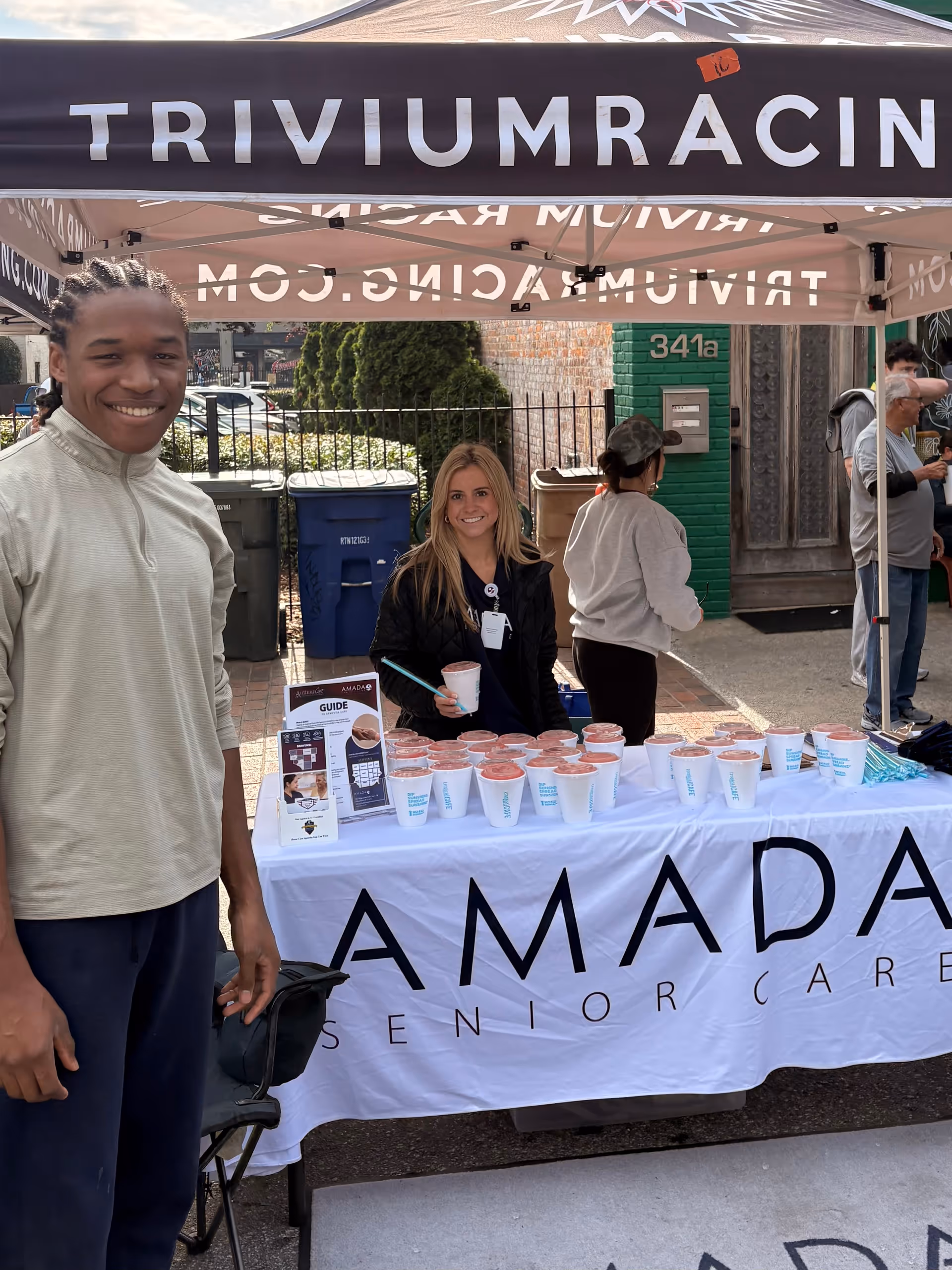Outdoor Amada Senior Care booth with staff and cups on a table under a tent.