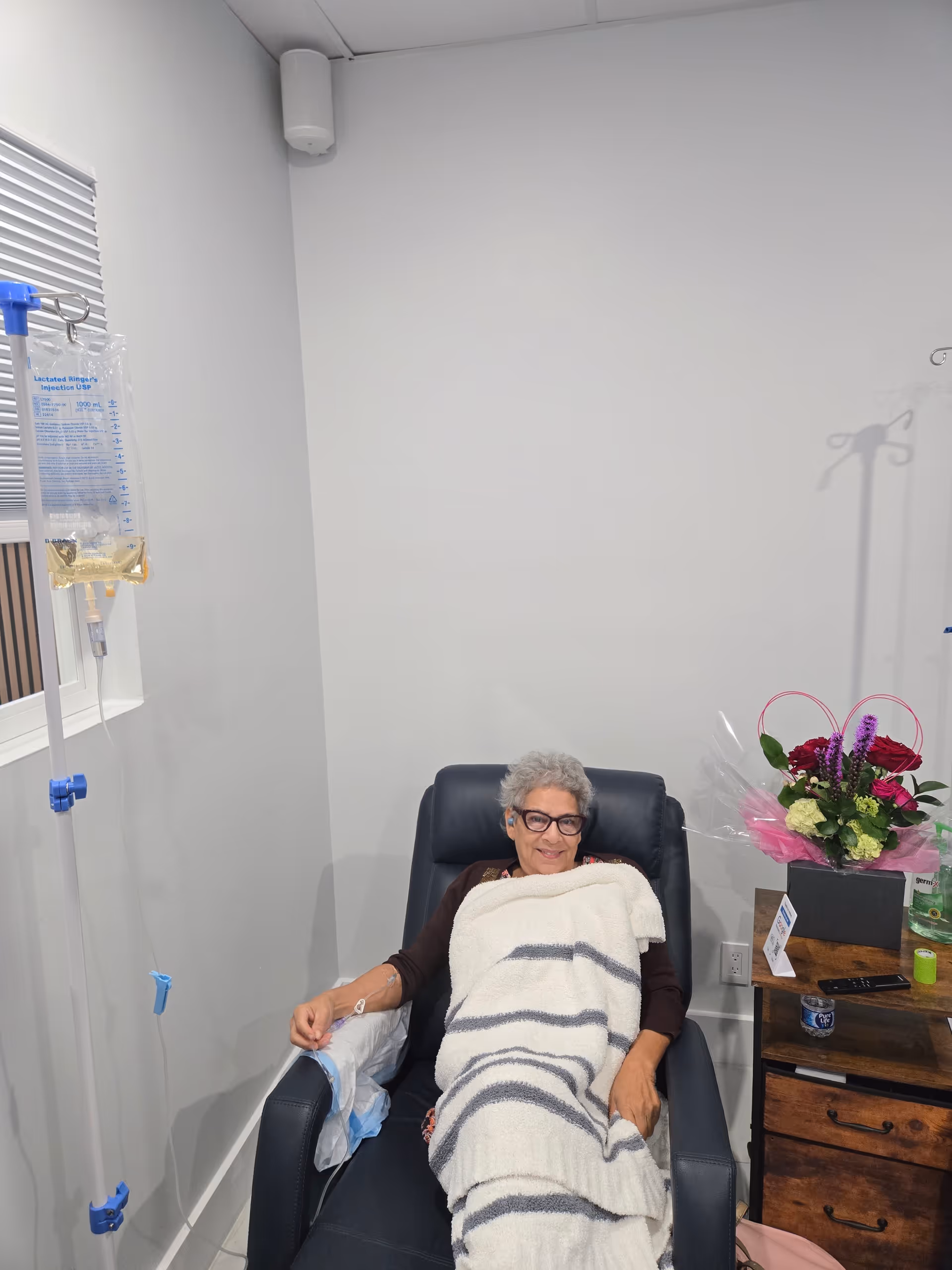 Elderly woman sits in an armchair with a blanket and IV bag in a medical room.