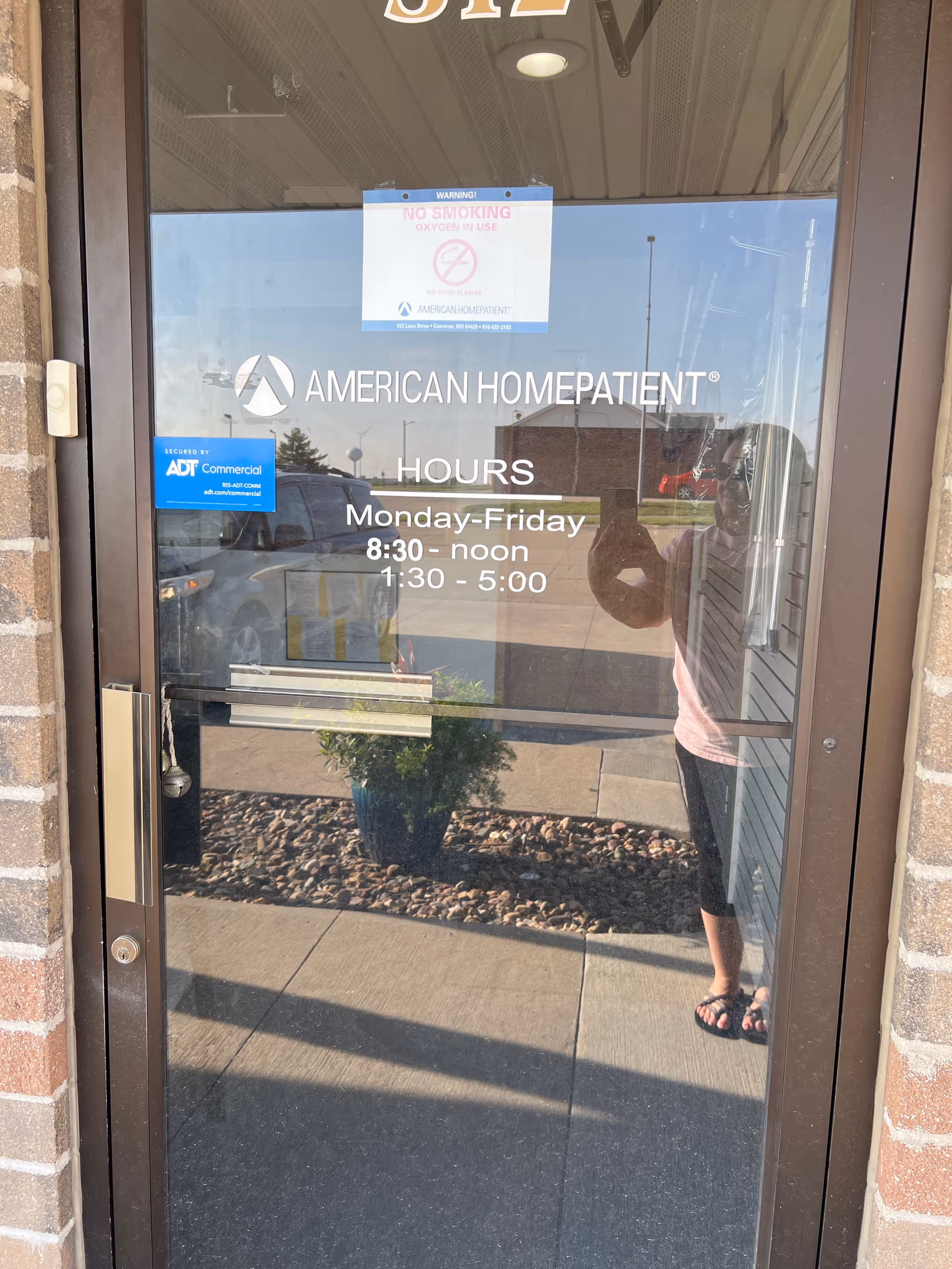 Glass entrance of American HomePatient with hours sign and a reflected person