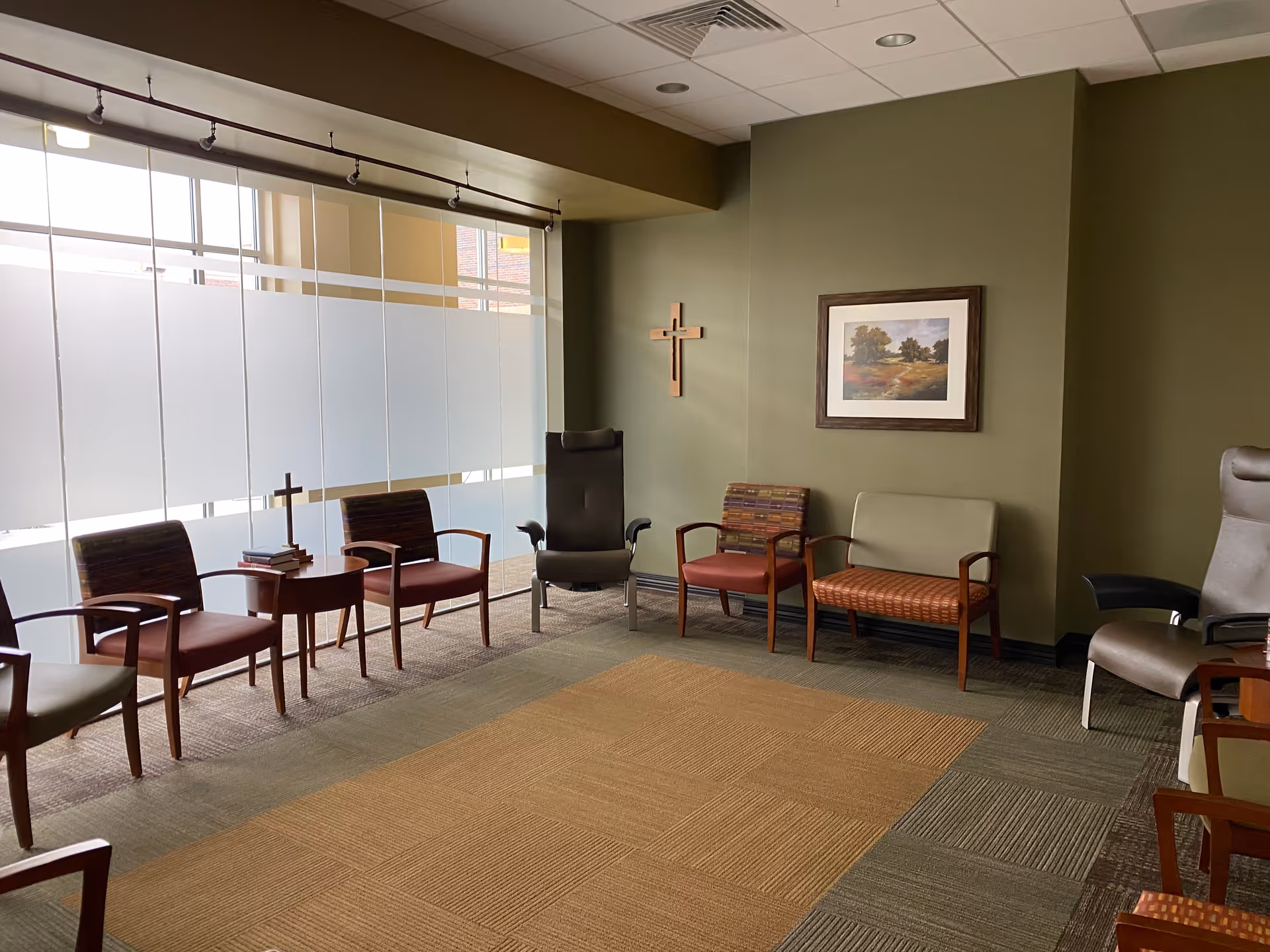 A quiet senior living lounge with chairs along frosted glass windows and a wall crucifix.