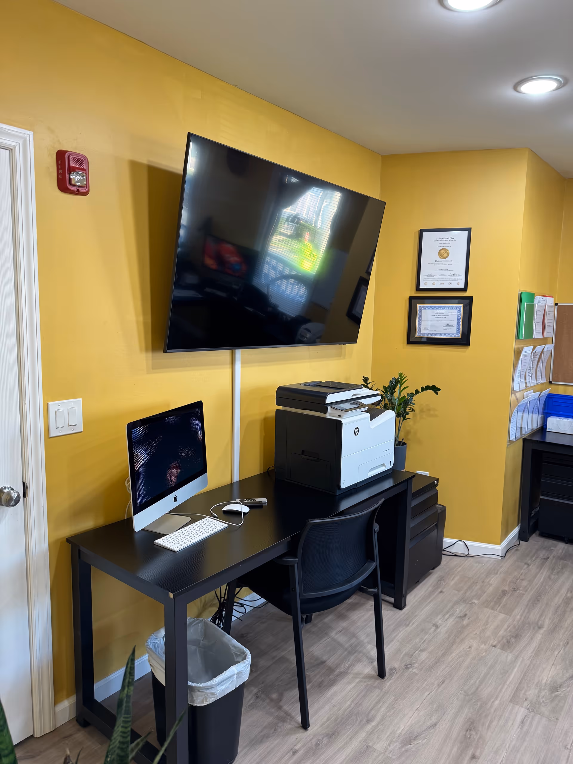 Yellow-walled office area with a desk, computer, printer, and a large wall-mounted TV.
