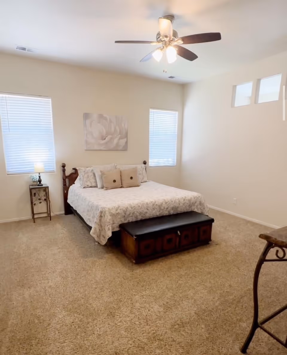 A neatly made bedroom with a bed, ceiling fan, and two windows.