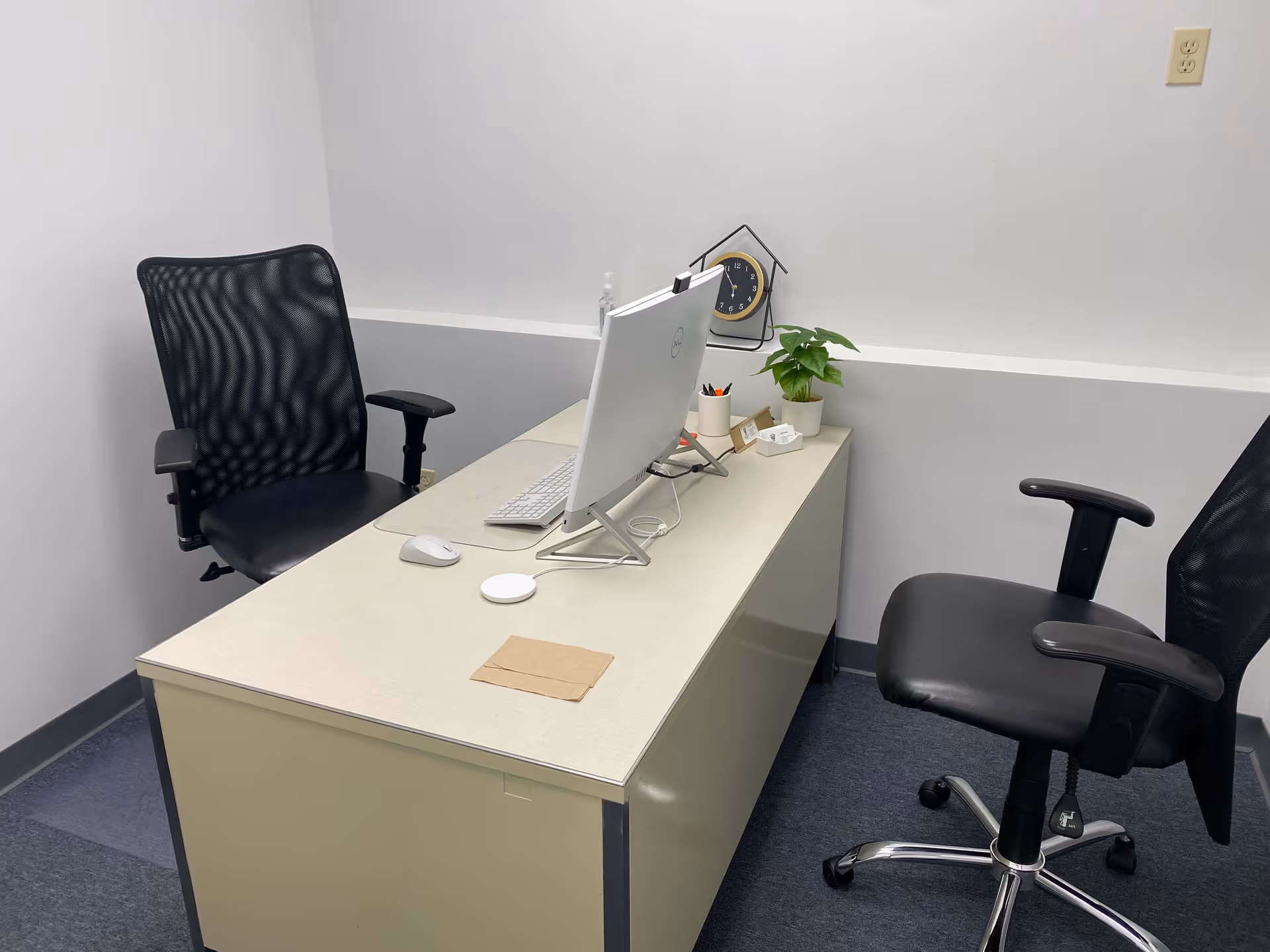 A neat desk setup with computer, two chairs, plant, and clock in an office.