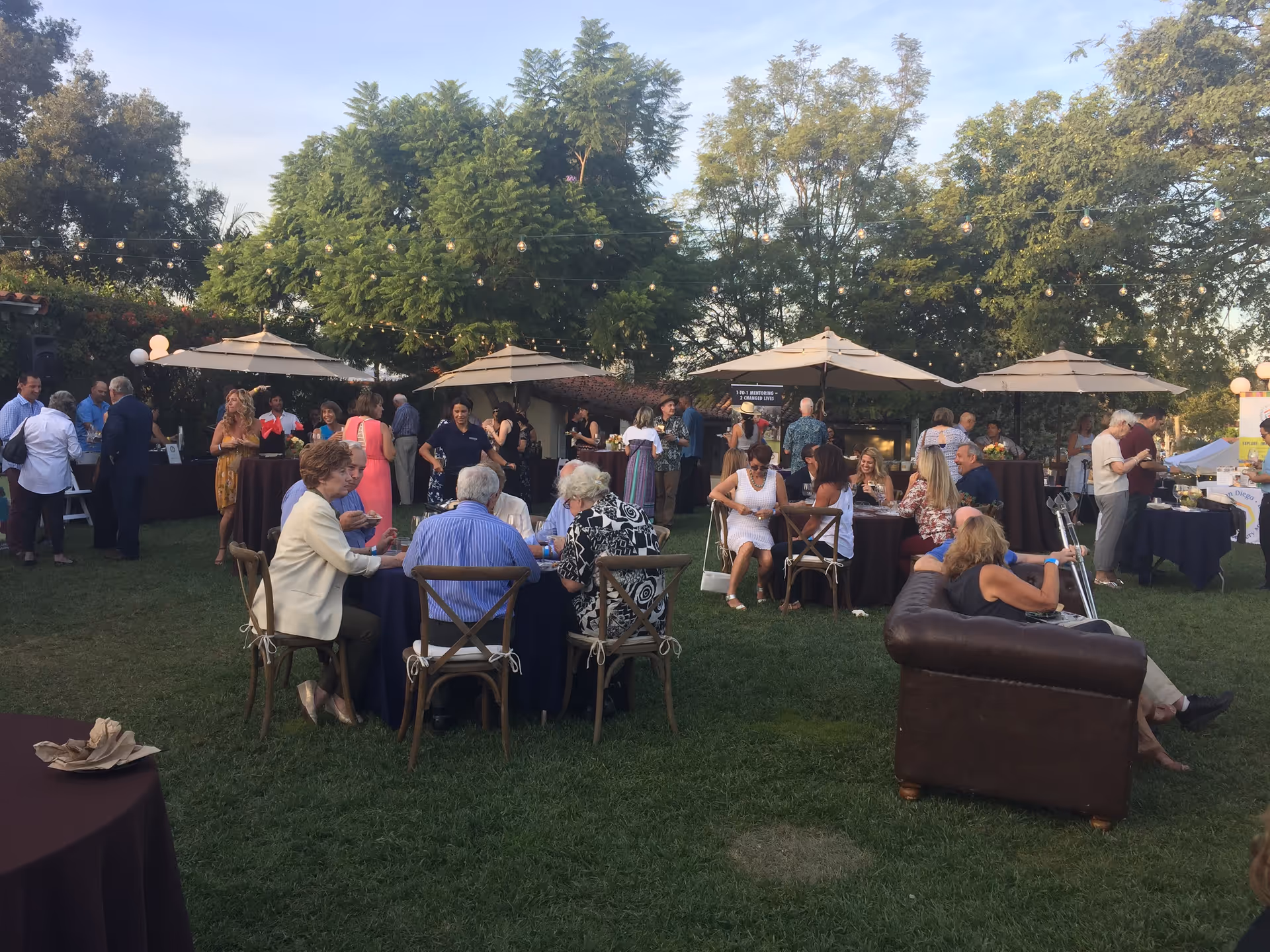 Outdoor garden event with guests seated at tables under umbrellas and string lights.