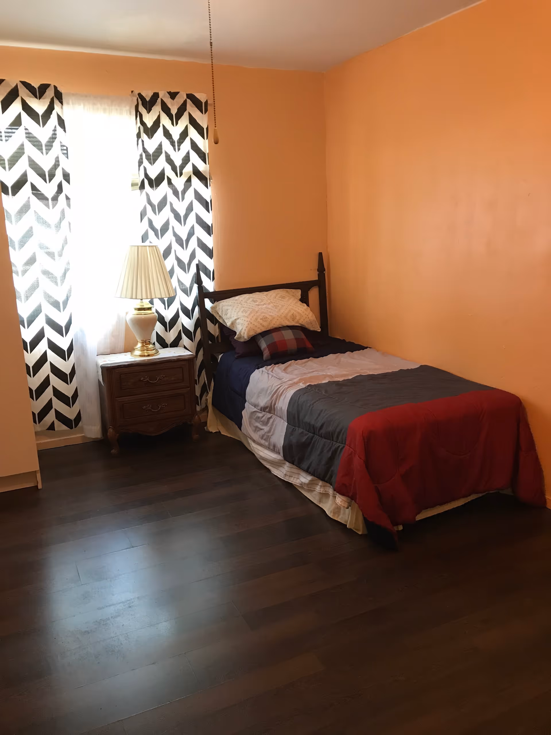 Small bedroom with a single bed, nightstand, lamp, and patterned curtains.