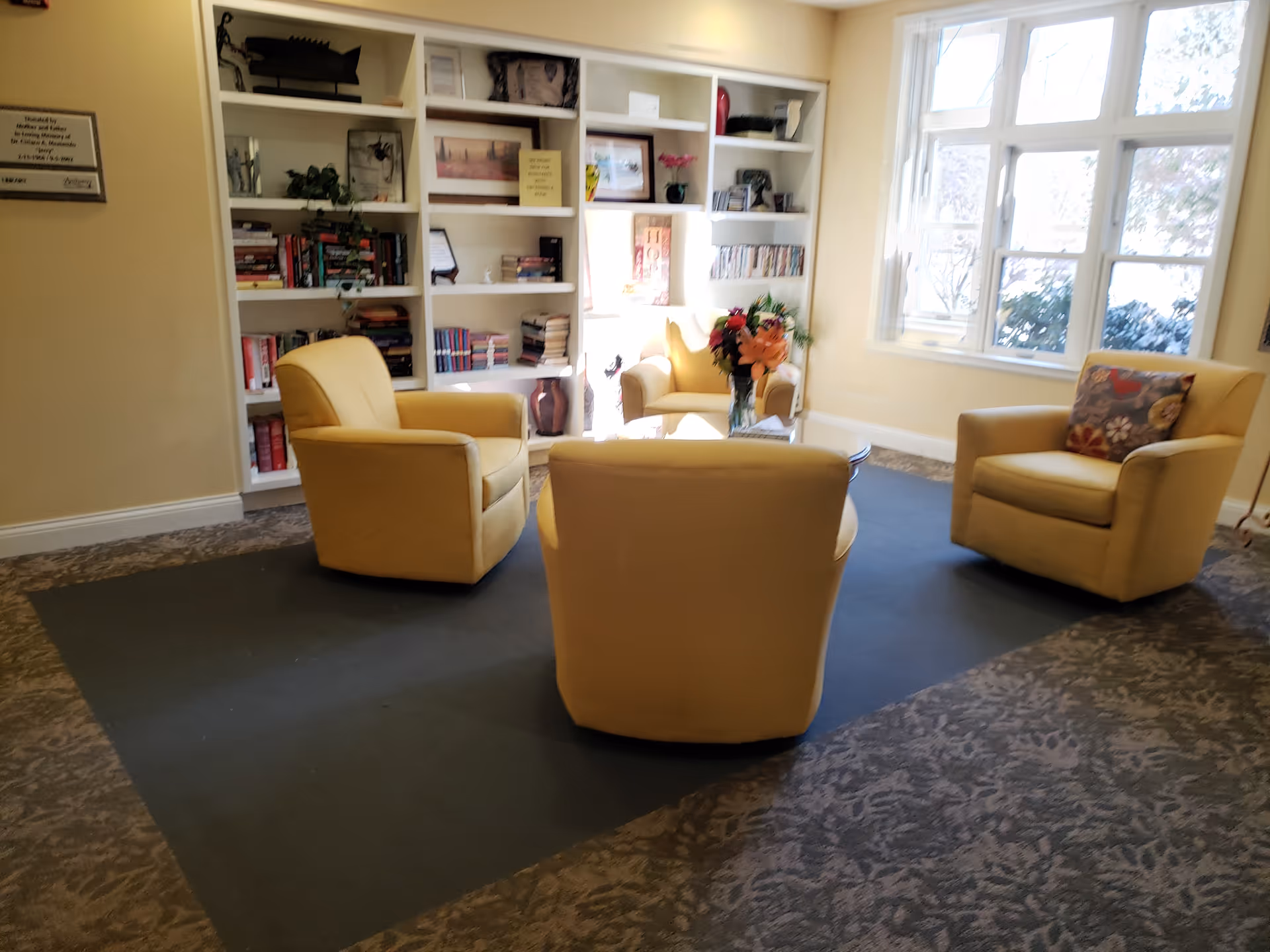 A cozy lounge in Mercy Hospice with beige chairs, a bookshelf wall, and a large window.