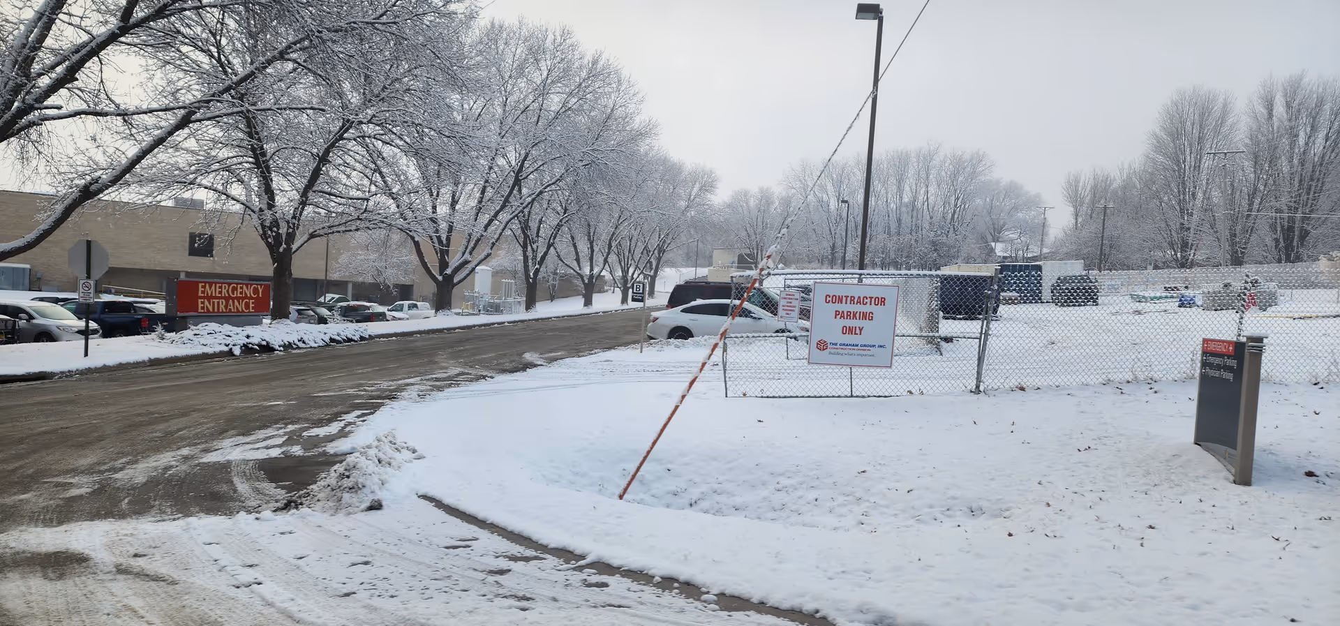 Snow-covered hospital entrance area with parked cars, signage, and chain-link fence.