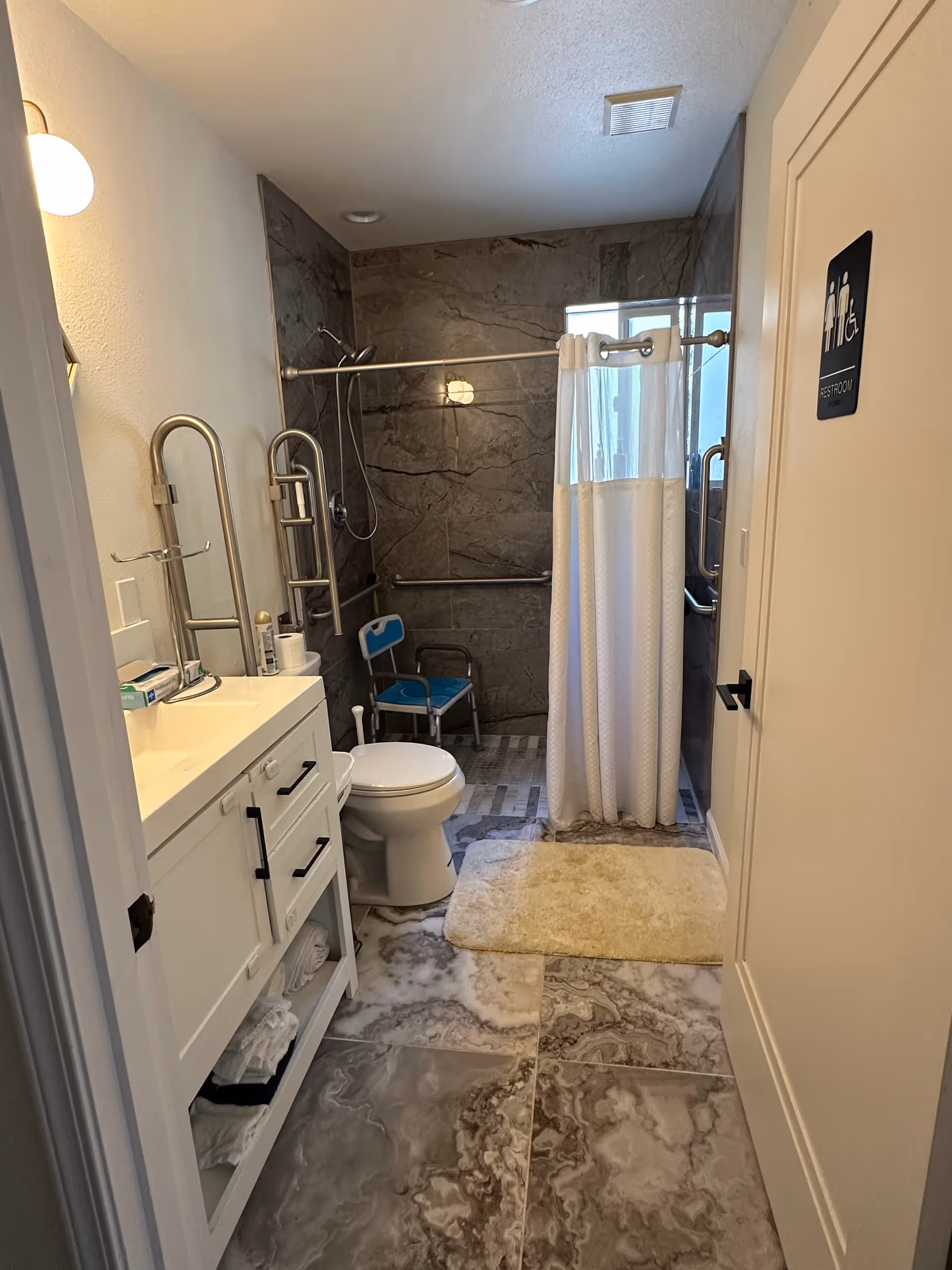 Accessible bathroom with sink, toilet, marble floor, shower with curtain, and grab bars.