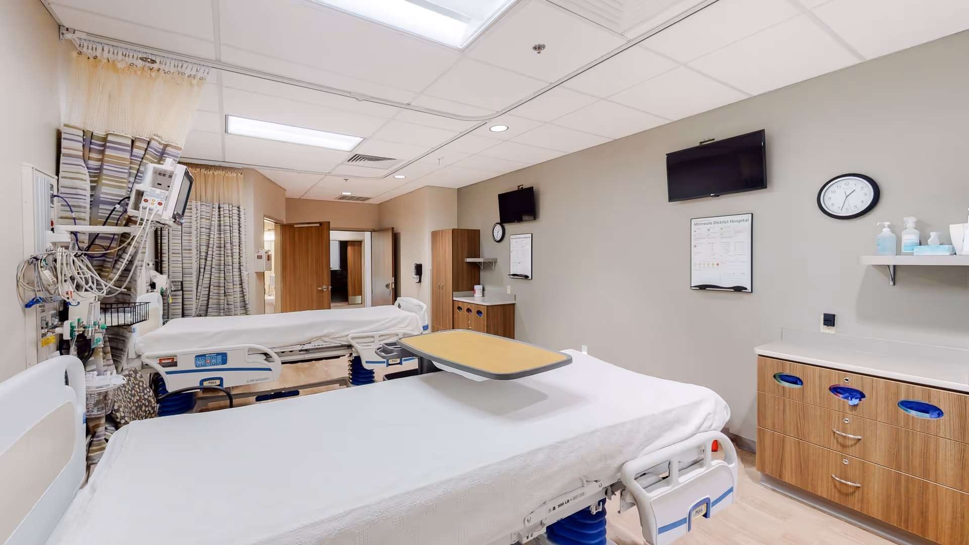 Interior hospital ward featuring two beds, medical equipment, curtains, monitors, and wall clock.