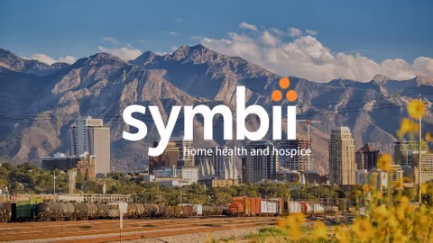 City skyline with mountains in background and yellow flowers in foreground; symbii watermark.