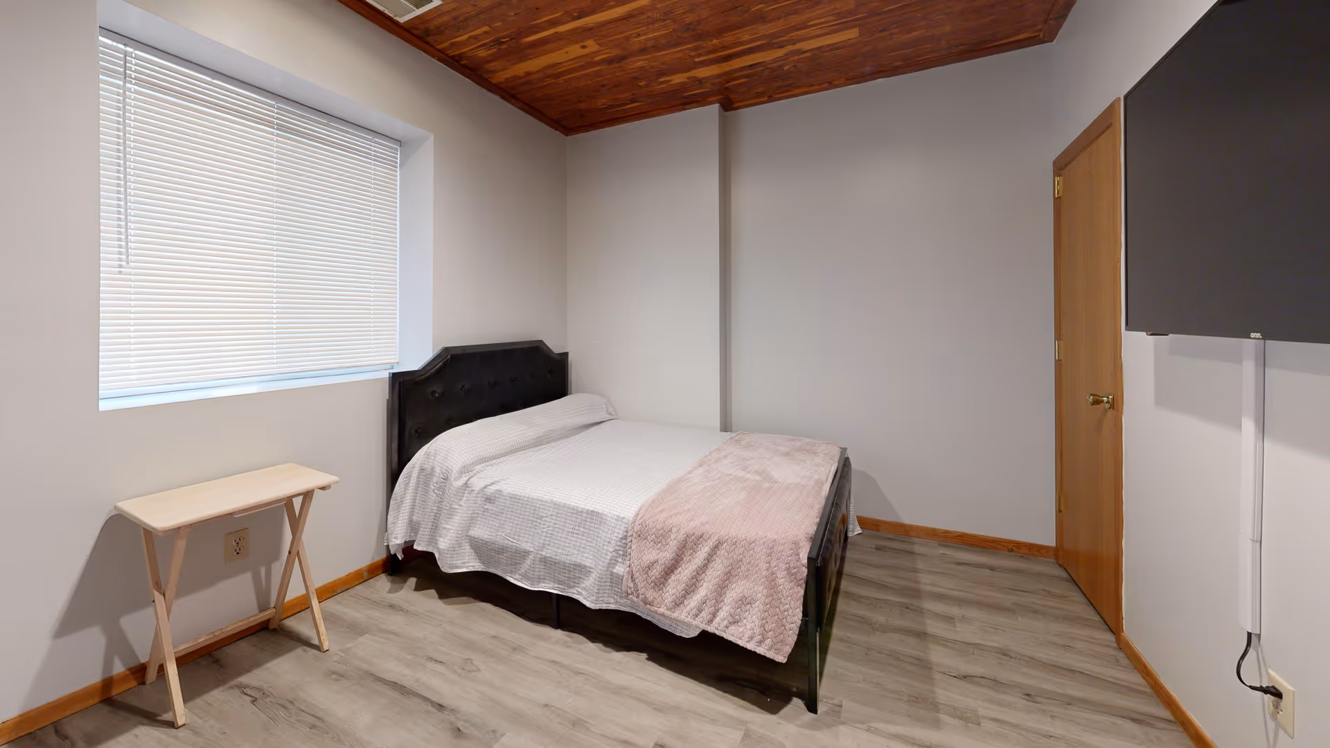 A small furnished bedroom with a bed, window blinds, a table, and a wall-mounted TV.