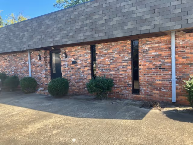 Brick facility front with a black doorway, windows, and trimmed shrubs.
