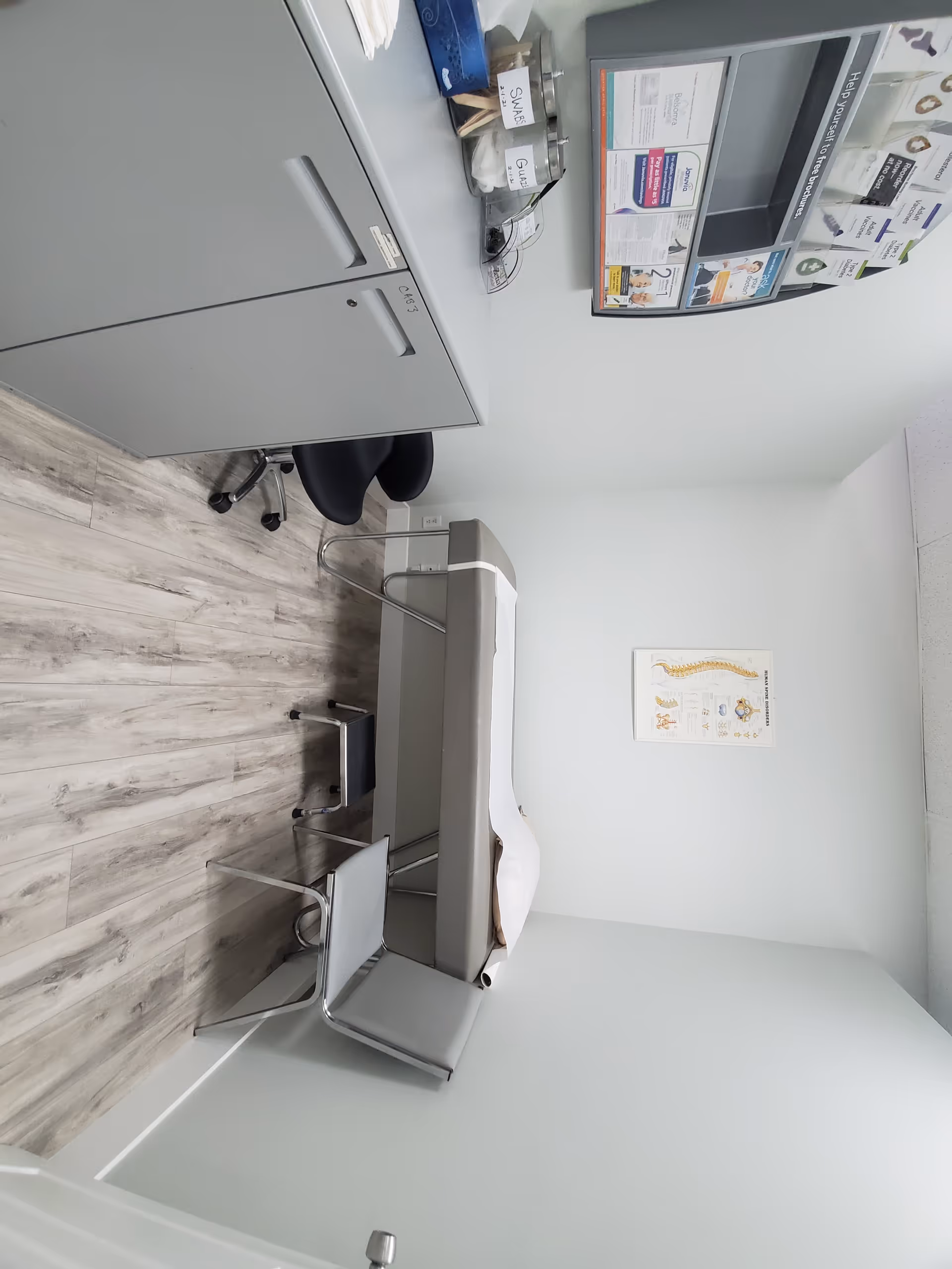 Clinic examination room with an exam bed, cabinets, chairs, and wall posters.